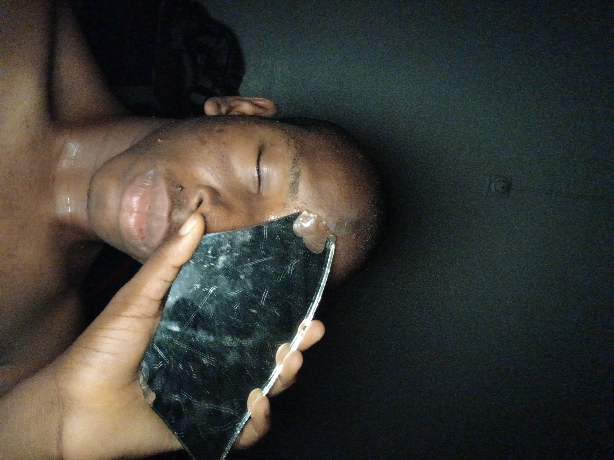 The image shows a close-up, dimly lit, vertical shot of a dark-skinned individual, likely male, from the chest up. The person appears to be lying down or positioned at an unusual angle, with their head resting on a dark surface. Their eyes are closed. Beads of sweat or liquid are visible on their forehead, cheek, and neck, and their lips are slightly parted.
A hand, belonging to the same individual or another person, is prominent in the foreground, holding a large, irregularly shaped piece of broken glass. The glass appears to be a fragment of a mirror or a phone screen, with visible cracks and scratches, reflecting some light. The sharp edge of the broken glass is held precariously close to the person's right eye and forehead.
The background is uniformly dark, suggesting either night-time or a very dimly lit indoor environment, possibly a room with dark walls or bedding. There are no discernible objects or features in the background apart from a faint, small, square shape with a circular element, possibly a wall fixture, in the upper right quadrant. No visible text is present in the image. The overall mood is somber and potentially unsettling due to the proximity of broken glass to the face. The location context indicates Lagos, Nigeria.
