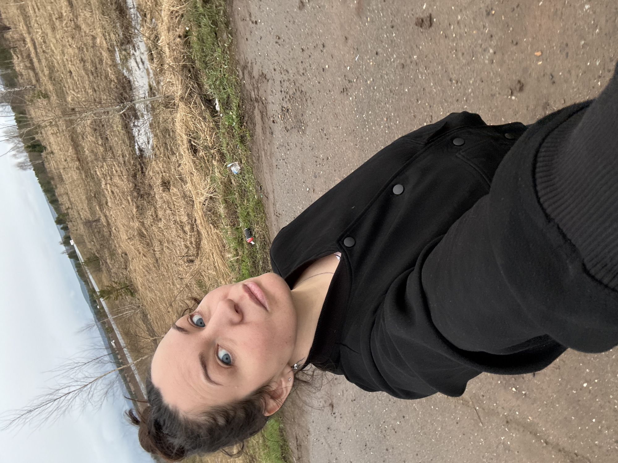 A selfie depicts a woman with light skin, blue eyes, and dark hair pulled back into a bun, wearing a black jacket and necklace. She stands outdoors on a dirt or gravel path. To her left is a field with patches of standing water and bare trees. The sky is overcast. The location is consistent with a rural or semi-rural area. Without any additional context or metadata, it is not possible to confirm or deny the specific location of Povarenki, Russia.