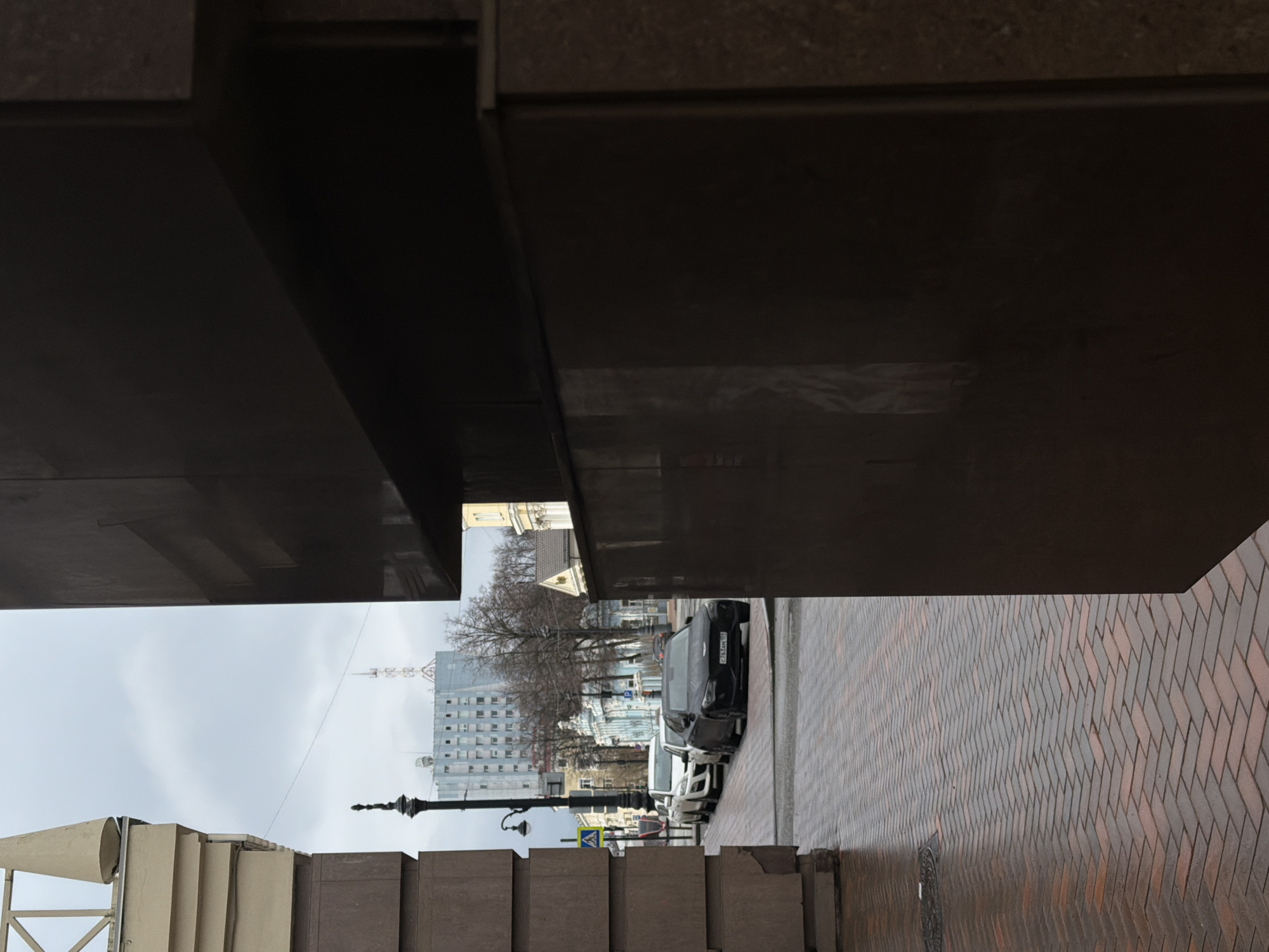 This image shows a street scene in Perm, Russia, viewed from under a dark-colored overhang. A herringbone pattern brick walkway is visible on the right. Cars are parked along the street to the left, and a tall building with a spire is in the background. A black lamppost is also visible. The sky is overcast.
