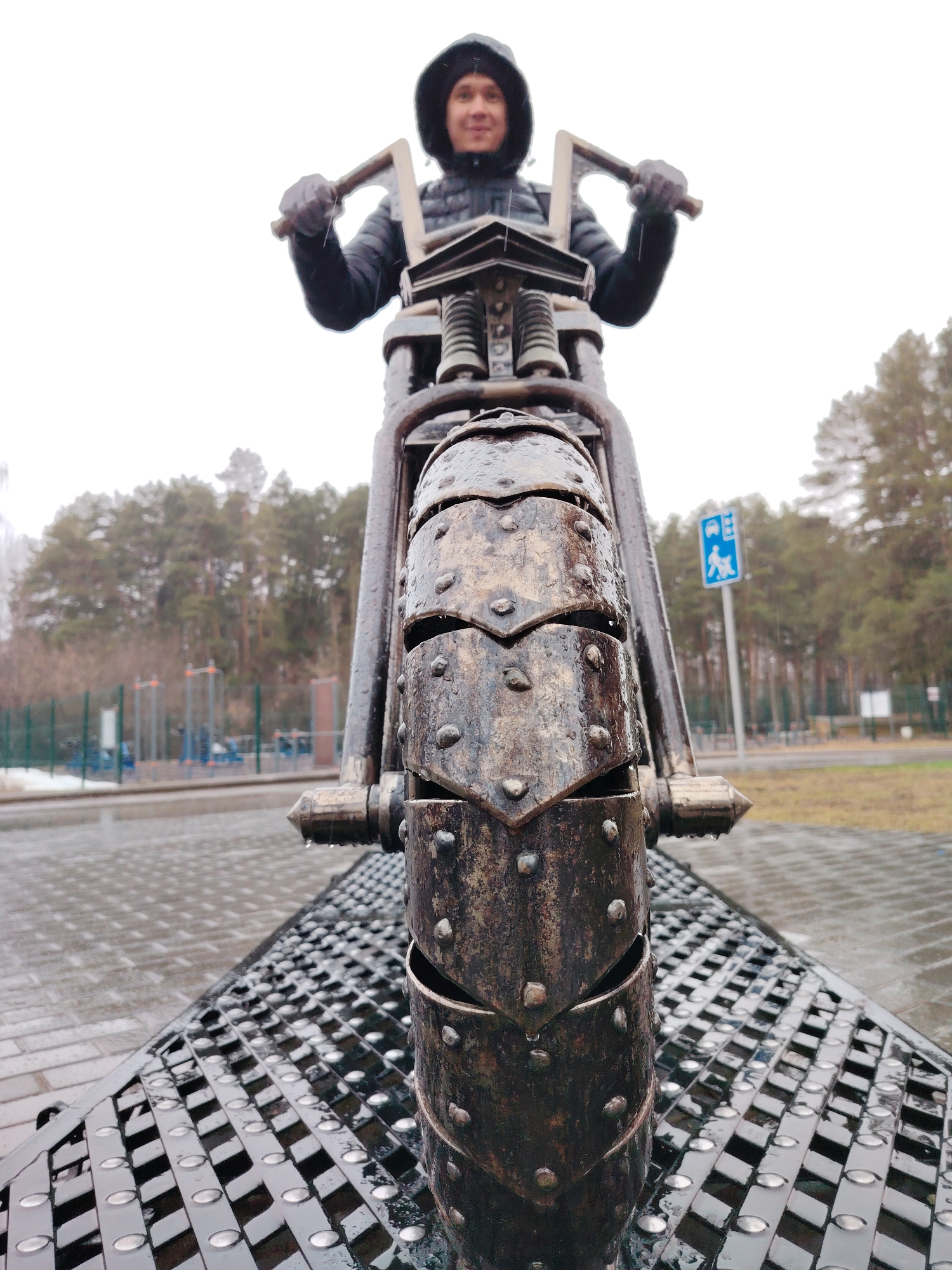 A low-angle, full shot captures a person on a metal motorcycle sculpture in Chaikovsky, Russia. The person, positioned behind the motorcycle's handlebars, wears a black jacket with a hood up and black gloves. The focus is sharply on the front wheel of the motorcycle, which is segmented and embellished with raised rivets. The motorcycle itself appears to be constructed from metal, with visible water droplets, implying recent or ongoing rain.
The surface under the motorcycle is a dark, metallic grid-like structure, also wet. In the background, out of focus, there is a park-like setting with bare trees, a blue and white public transportation sign, and a playground. The sky is overcast, creating diffused lighting. The ground is paved and wet, reflecting ambient light.