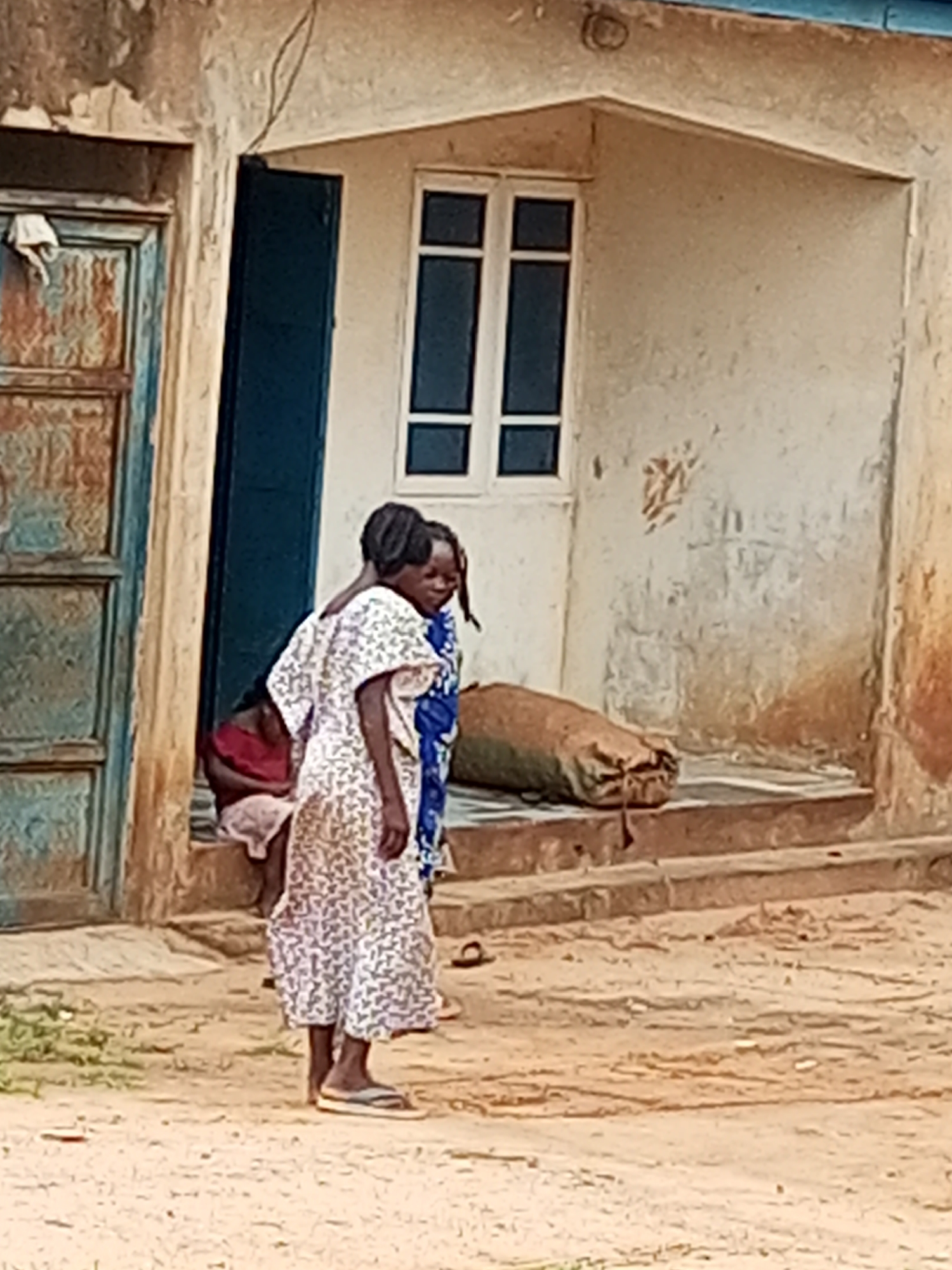 This is a medium shot, outdoors, full-color, eye-level photograph taken in Tempure, Nigeria. The scene depicts two young Black females standing on a dirt path in front of a weathered building. The building has a light-colored stucco exterior with a dark blue door to the left and a large window with white frames and dark panes in the center. The door appears aged and has visible rust. To the right of the door, a person in a red top and light-colored bottoms is partially visible, sitting on the steps. The female in the foreground on the left is wearing a long, white dress with a subtle floral pattern and appears to be looking towards the right. The second female, partially behind the first and to her right, is wearing a blue and white patterned garment and is facing away from the camera. Between the two females and slightly behind them, a large, cylindrical burlap sack lies on the steps. The ground is sandy dirt, and the overall environment suggests a rural or semi-urban setting in Nigeria.