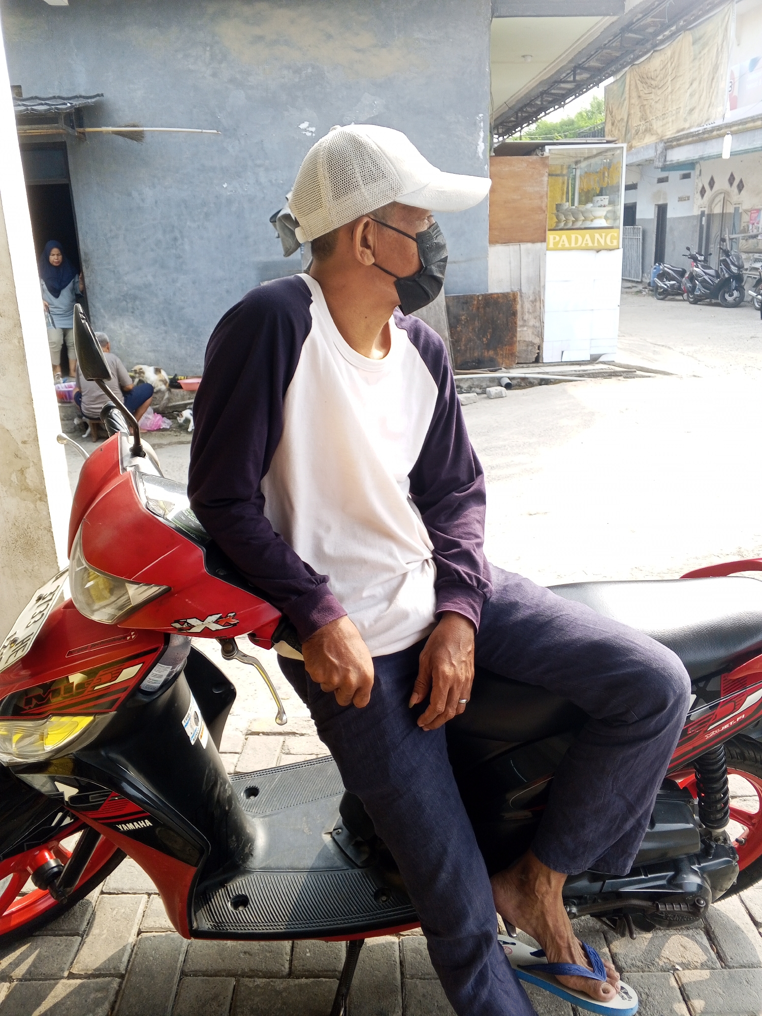 A man wearing a white baseball cap, a dark purple and white long-sleeved shirt, dark blue pants, and blue flip-flops sits on a red and black Yamaha scooter. He is wearing a black face mask and a ring on his left hand. He is looking to his left. The scooter is parked on a paved area in front of a building. In the background, a woman in a blue headscarf stands in a doorway. There are several motorcycles parked further in the background. A sign that reads "PADANG" is visible on a glass-fronted kiosk. The scene appears to be an outdoor area in a city.