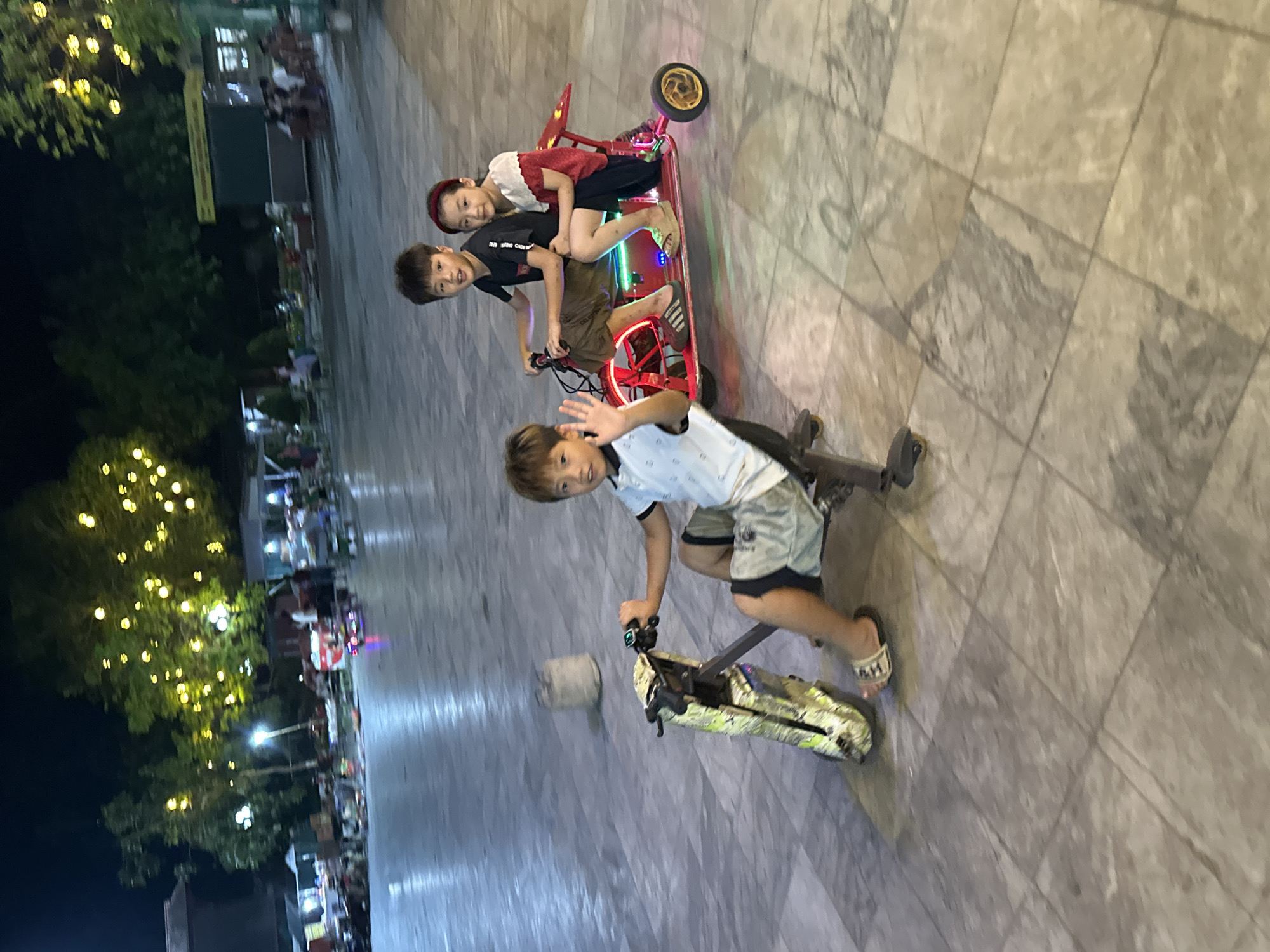 This image captures three children enjoying electric ride-on toys outdoors at night in Phố Ràng, Vietnam. In the foreground, a boy with light brown hair, wearing a white polo shirt, light-colored shorts, and H&M sandals, rides an electric scooter or tricycle. He is smiling and waving towards the viewer.
Behind him, two other children – a boy and a girl – share a larger, red electric ride-on toy, possibly a drift trike or go-kart, which is illuminated by internal green and blue LED lights. The girl, dressed in a red and white top, sits in front, while the boy, wearing a dark t-shirt and shorts, appears to be steering from the rear. Both children on the red vehicle also look happy and engaged.
The setting is an outdoor, paved area, likely a public square or recreational space. The time is nighttime, indicated by the dark background and the presence of numerous bright yellow string lights decorating trees in the left background, creating a festive ambiance. Distant, blurry lights and structures suggest other activity or stalls in the background. The children appear to be of Asian descent. The scene conveys a sense of fun and leisure as the children enjoy their evening rides.