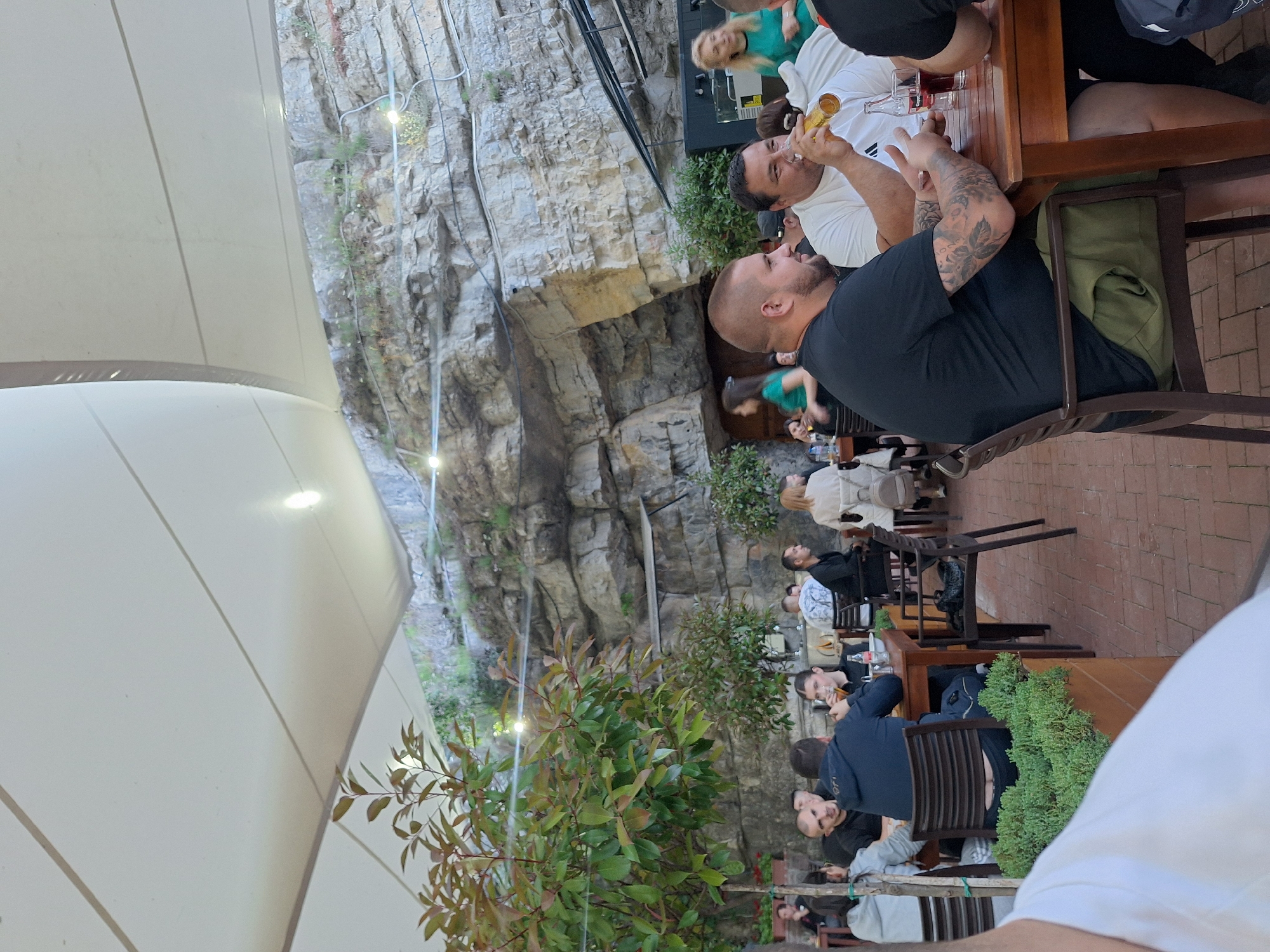 The scene is an outdoor restaurant with patrons seated at wooden tables. The restaurant is constructed at the base of a rock cliff face, with greenery interspersed. People are eating and drinking. The location is Milanovo, Bulgaria.