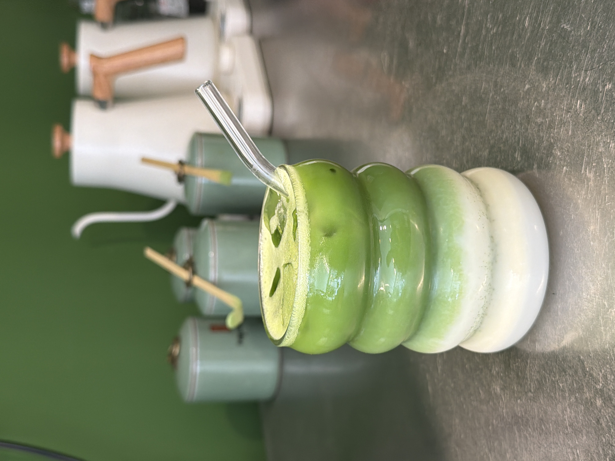 In a close-up shot, a layered green and white beverage sits in a uniquely shaped glass with a glass straw. The drink, likely a matcha latte, displays a creamy white base and transitions to a vibrant green at the top, topped with ice cubes and a few leafy garnishes. The glass itself has a ribbed, bulbous design, adding a modern aesthetic to the drink. In the background, out of focus, are several green storage containers with gold accents, and two white kettles with wooden handles, suggesting a cafe or kitchen setting. The overall scene is clean and brightly lit, with a focus on the refreshing drink.
