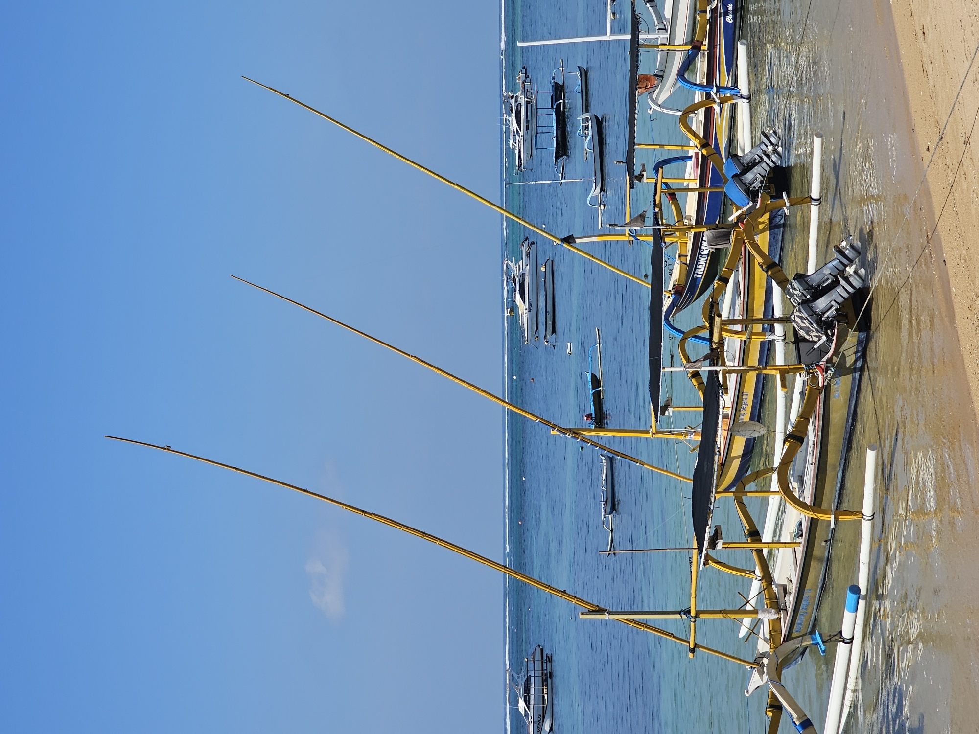 The image depicts a tranquil scene at a beach in Denpasar, Indonesia, likely Sanur Beach given the characteristic outrigger boats. Several traditional Balinese jukung boats, identifiable by their yellow bamboo outriggers and outboards, are moored in the shallow, clear blue waters close to the sandy shore. The boats are arranged in a line, some appearing to be pulled up onto the sand. The water is calm, with gentle waves lapping at the beach. In the background, a few white yachts are anchored further out, and the horizon is marked by a thin white line of breaking waves. The sky is a vibrant, cloudless blue, suggesting a sunny day. The scene is devoid of any visible people or immediate activity, conveying a sense of peacefulness and waiting. The overall impression is one of a picturesque coastal landscape in Denpasar, ready for the day's activities.