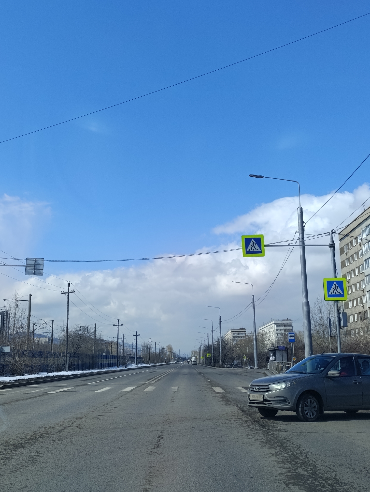 The image shows a wide, straight road in Krasnoyarsk, Russia. A gray car is parked on the right side of the road. Two pedestrian crossing signs are visible next to the car, attached to a lamppost. In the distance, there is a bus stop and residential buildings. On the left side of the road are railroad tracks. Overhead power lines stretch across the sky, which is mostly blue with some clouds. Patches of snow remain on the roadside.
