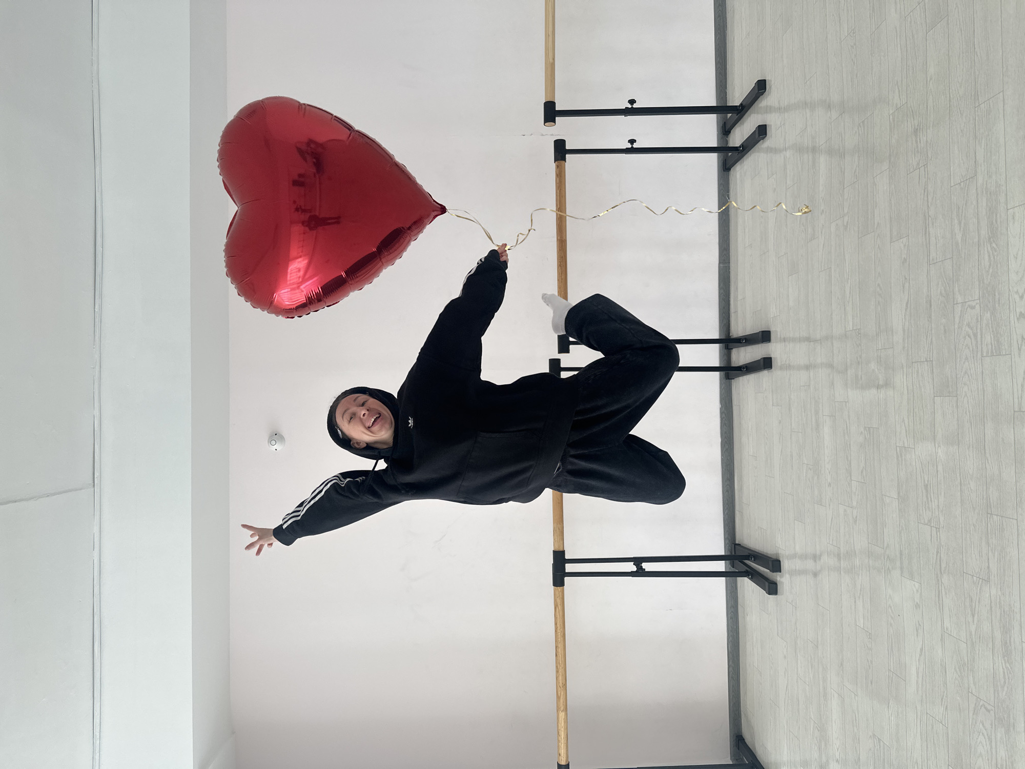 A person in a black Adidas hoodie and jeans is suspended upside down, holding a red heart-shaped balloon, in what appears to be a dance studio. The room has white walls and a light gray wood-patterned floor. There are two ballet barres visible, one with a wooden bar and one with black metal bars. The photograph may have been taken in Votkinsk, Russia.