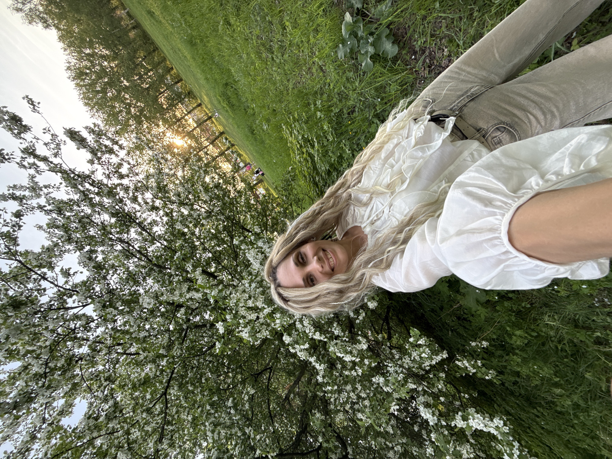 The image shows a woman with long, blond hair wearing a white blouse and beige pants, lying in a grassy area in Шебунино, Russia. A tree with white blossoms fills the lower left portion of the frame. The background consists of green grass and trees, with a hint of sunlight filtering through the foliage in the upper-left corner.