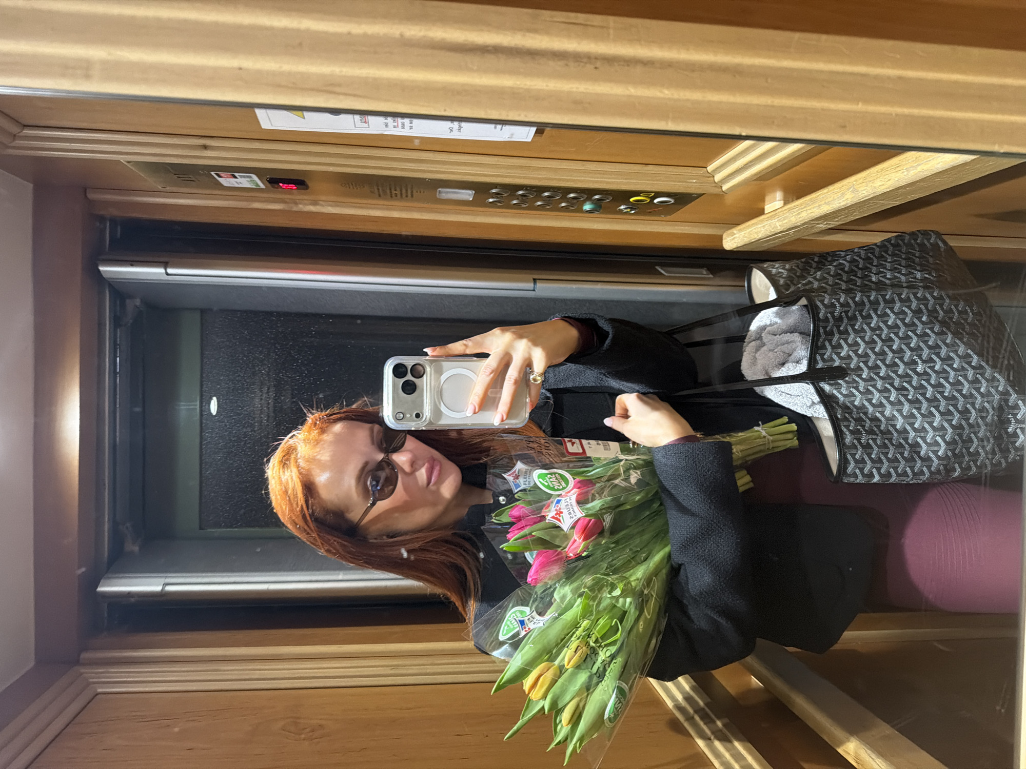 A person with red hair is taking a selfie in an elevator, likely in Paris, France, based on the style and design of the elevator and bag. They are holding a bouquet of wrapped tulips. The elevator has wooden paneling and the button panel is visible above. A gray patterned bag and a purple seat are also visible in the elevator.