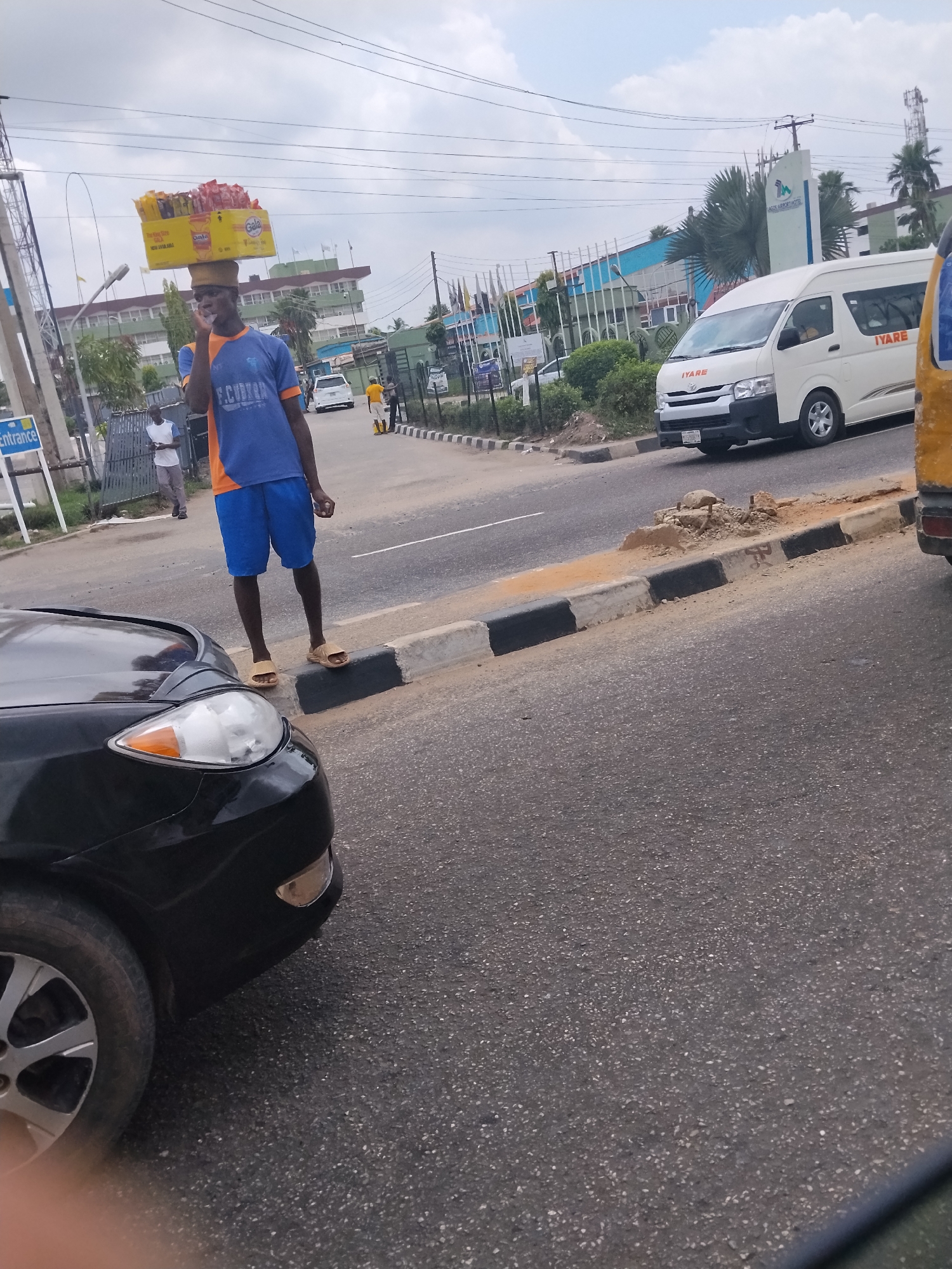 This photograph captures a bustling street scene in Ikeja, Nigeria, under an overcast daytime sky. The central figure is a young man, likely a street vendor, standing on a black and white painted curb alongside a busy road. He is dressed in a blue and orange short-sleeved shirt with "F.C. CUSTOM" printed on the chest, blue shorts, and light brown sandals. He skillfully balances a large yellow cardboard box, filled with snack items such as "Gala" branded sausage rolls, on his head. He appears to be holding a phone to his ear, possibly engaged in a conversation.
In the background, a white Toyota Hiace passenger van, bearing the word "IYARE" on its side, is visible on the road. Further back, a building complex is prominent, marked by a vertical sign that reads "LAGOS AIRPORT HOTEL," providing a clear location cue for Ikeja. Power lines crisscross the sky above. In the foreground on the left, the hood, headlight, and tire of a black car are visible, suggesting the photo was taken from a vehicle. The road surface is asphalt, and there are areas of dirt and minor construction debris along the curb. A sign partially visible on the far left indicates an "Entrance." The image depicts a common aspect of informal commerce and urban life in Nigeria.