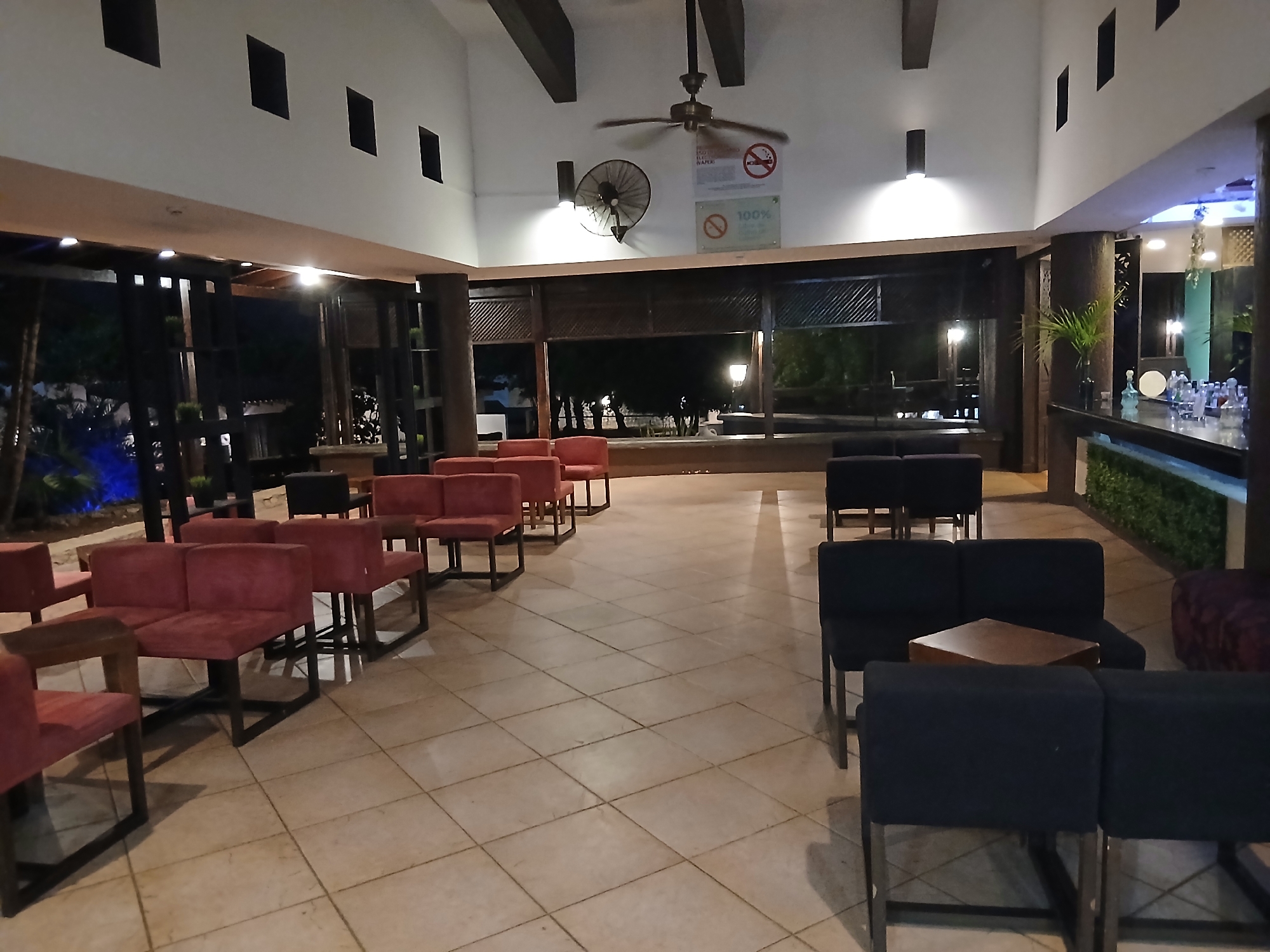 The image depicts an indoor seating area in what appears to be a bar or lounge. The location is specified as Playa El Agua, Venezuela. A variety of seating options are visible, including red upholstered chairs and couches and dark blue upholstered chairs. Some of the chairs are arranged around small wooden tables. The floor is tiled. The ceiling is high, with visible wooden beams and a ceiling fan. A wall fan is visible. Signage for "100% free" and "no smoking" is also visible near the ceiling. The area seems open to an outdoor space, as evidenced by the view through the large openings revealing plants, lights, and possible beach scenery. To the right, there is a bar area with a counter and various items displayed on it. No people are clearly visible in the frame.