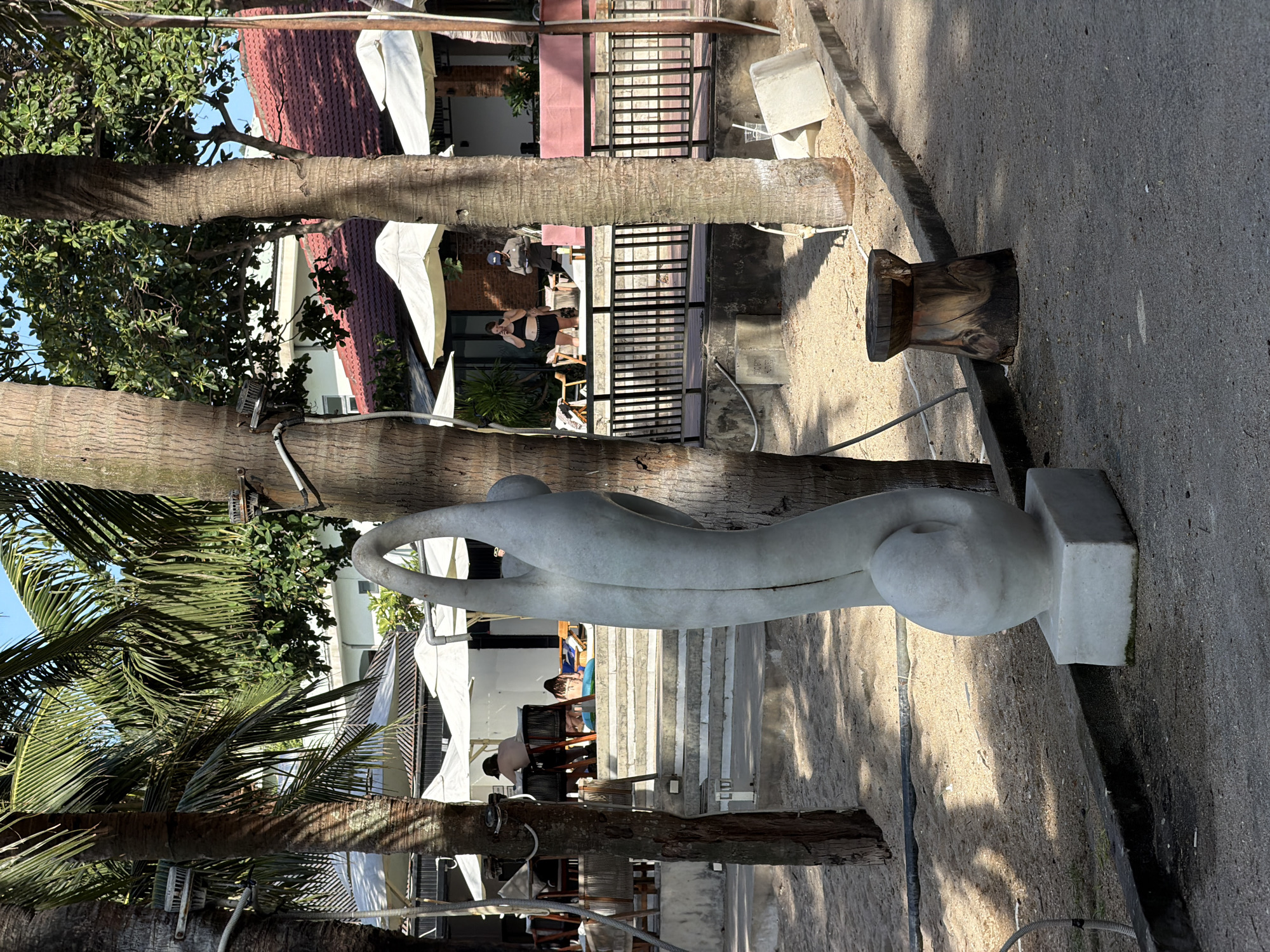 The image depicts a beachside restaurant or resort setting, likely in the Mỹ Khê Beach area of Đà Nẵng, Vietnam, given the presence of palm trees, beach sand, and a building with a tiled roof. In the foreground, a modern white abstract sculpture of a human form stands on a square base. To its right is a rustic wooden stool. The background reveals a shaded seating area with white umbrellas, tables, and chairs. Several people are visible, some seated at tables, others reclining on lounge chairs or on steps leading up to the seating area. One person is standing near a table, holding a drink. Another individual, wearing a cap, appears to be working or serving. The environment is sandy with paved walkways and concrete structures. The lighting suggests it is daytime, with shadows cast by the palm trees.