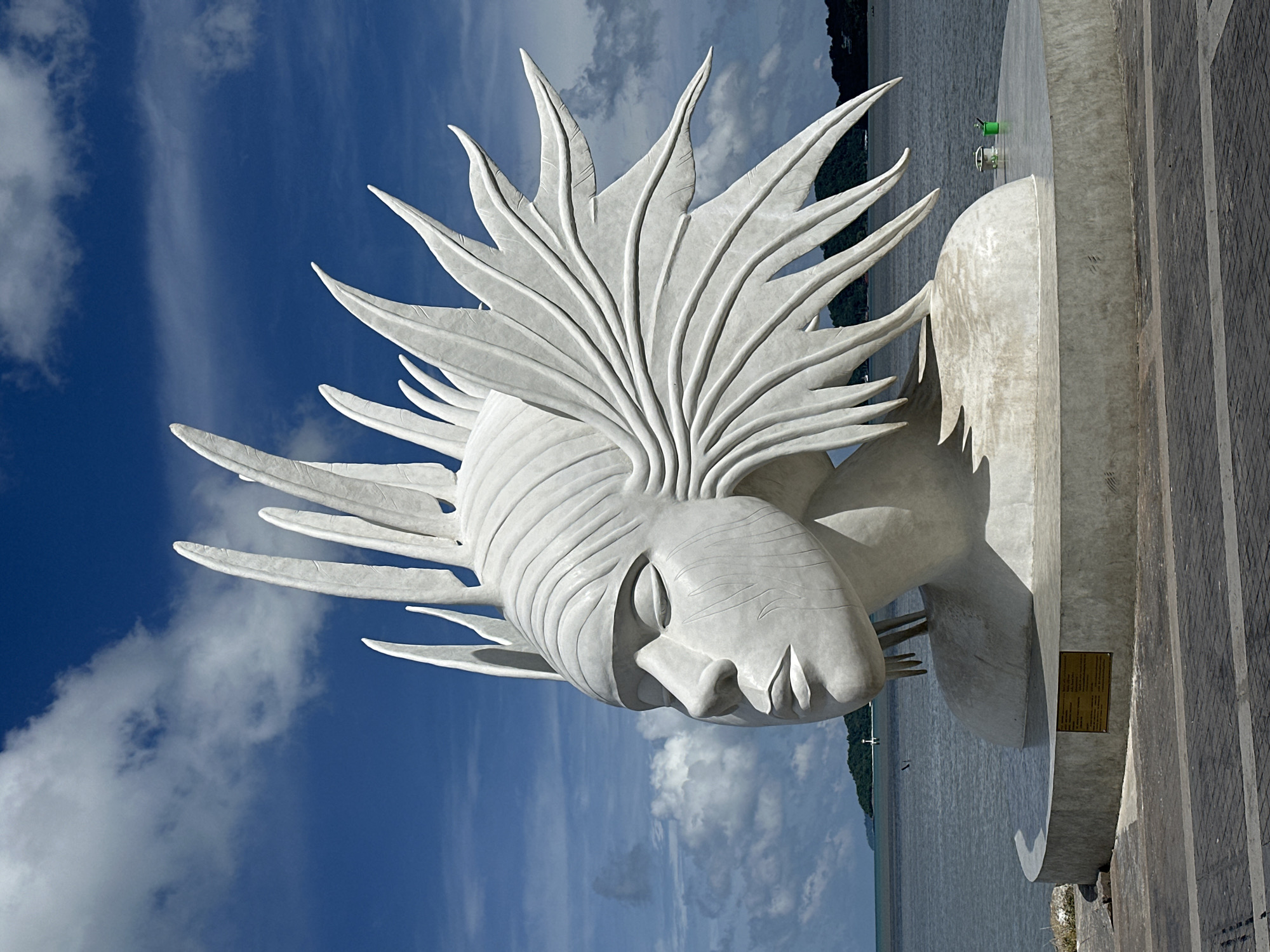 A large, white, concrete sculpture of a woman's head and shoulders stands by the sea under a bright blue sky with scattered clouds. The sculpture depicts a serene, closed-eyed face with stylized, feather-like headdress. The base of the sculpture is circular and made of concrete. A plaque is affixed to the front of the base. The sculpture is situated on a paved promenade. In the background, the sea stretches to the horizon, with a distant island and lighthouse visible. Two green buckets and some brushes are on the concrete platform to the right of the sculpture. The scene appears to be in Phuket City Municipality, Thailand, possibly along the waterfront promenade.