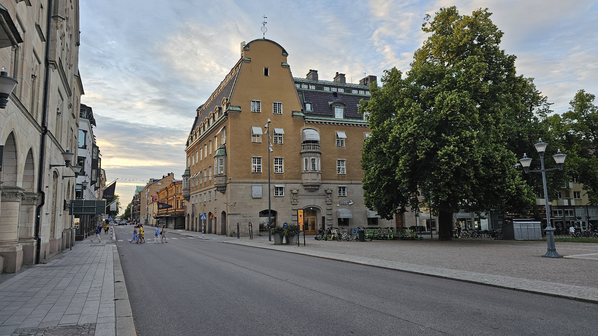 The image depicts a street scene in Linköping, Sweden, specifically on Sankt Larsgatan, near the intersection with Trädgårdstorget. The perspective is from the middle of the street looking towards the east.
Dominating the left side of the frame is a multi-story building with arched ground-floor entrances and classical architectural details. This building is likely the Swedish Tax Agency (Skatteverket) building, characterized by its stone facade and ornate columns. Adjacent to it, further down the street, are other buildings with varied architectural styles, including a prominent yellow building with a distinctive mansard roof featuring dormer windows. This yellow building, with its rounded bay window and decorative stonework, is a focal point.
On the right side of the street, a large, mature tree with lush green foliage occupies a significant portion of the frame, casting shadows onto the paved area and the road. A streetlamp with multiple globes stands in the foreground on the right. Beyond the tree, other buildings are visible, including those lining Trädgårdstorget, which appear to house shops and businesses. Some of these facades have awnings and large windows.
Several people are present. On the sidewalk to the left, a few individuals are walking or standing. In the middle of the street, at a crosswalk, a group of people are crossing. Some are wearing bright yellow shirts. The road is asphalt, with clear lane markings. The sidewalk on the left is paved with light-colored tiles.
In the background, the street continues to recede, lined with more buildings and trees. The sky above is a mix of blue and white clouds, with soft, diffused light suggesting either early morning or late afternoon. A few electric scooters are parked on the paved area to the right of the yellow building, and a rack of bicycles is also visible. The overall atmosphere is calm and urban, with elements of historical architecture and modern amenities.