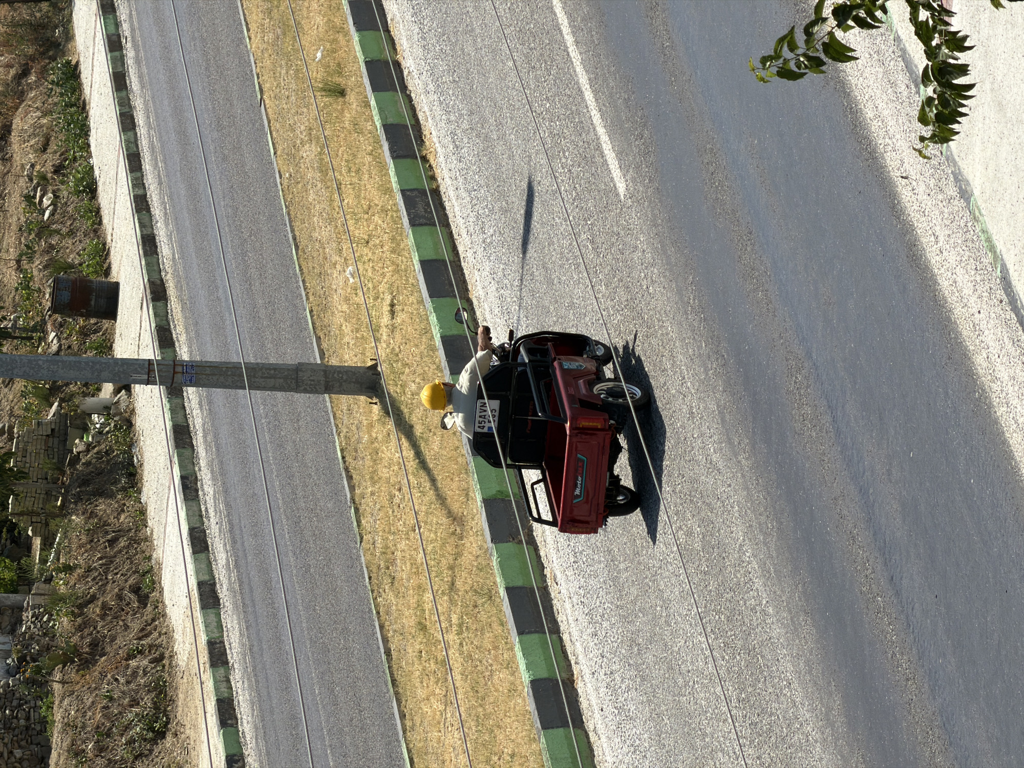 An overhead, medium shot captures a person wearing a yellow hard hat riding a small, red, three-wheeled vehicle on a paved road. The vehicle has a black cargo bed at the rear and the person is seated in front, facing away from the camera. The vehicle has a license plate that reads "45 AVN 595". The road is bordered by a grassy area on the left and a curb with alternating green and black painted blocks. In the background, a dirt embankment with sparse vegetation and some stone structures is visible. The scene appears to be on the outskirts of Köprübaşı, Turkey, judging by the general landscape and style of the roadside features. The lighting suggests it is daytime, with the sun casting a shadow from the vehicle onto the road.