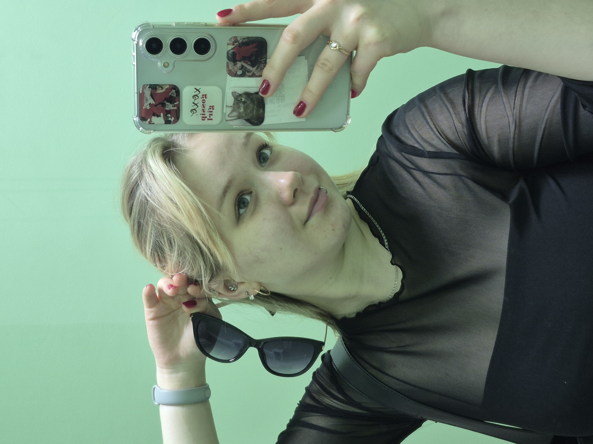 A light-skinned, young woman is taking a selfie in front of a mint green wall. She is holding a cell phone in her right hand above her head and angling the camera toward her face. Her left hand is holding a pair of black sunglasses, which she is using as a prop in the photo. The woman has light blonde hair, blue eyes, and is wearing a black mesh top. She has multiple piercings in her left ear and a small piercing above her lower lip on the left side. The cell phone has a clear case with stickers on the back including a cat, a graphic of "gossip girl xoxo" and other stickers. The woman is wearing a gold ring on her right ring finger. The photo appears to be taken indoors. The location of Chaikovsky, Russia, is not discernible from the image.