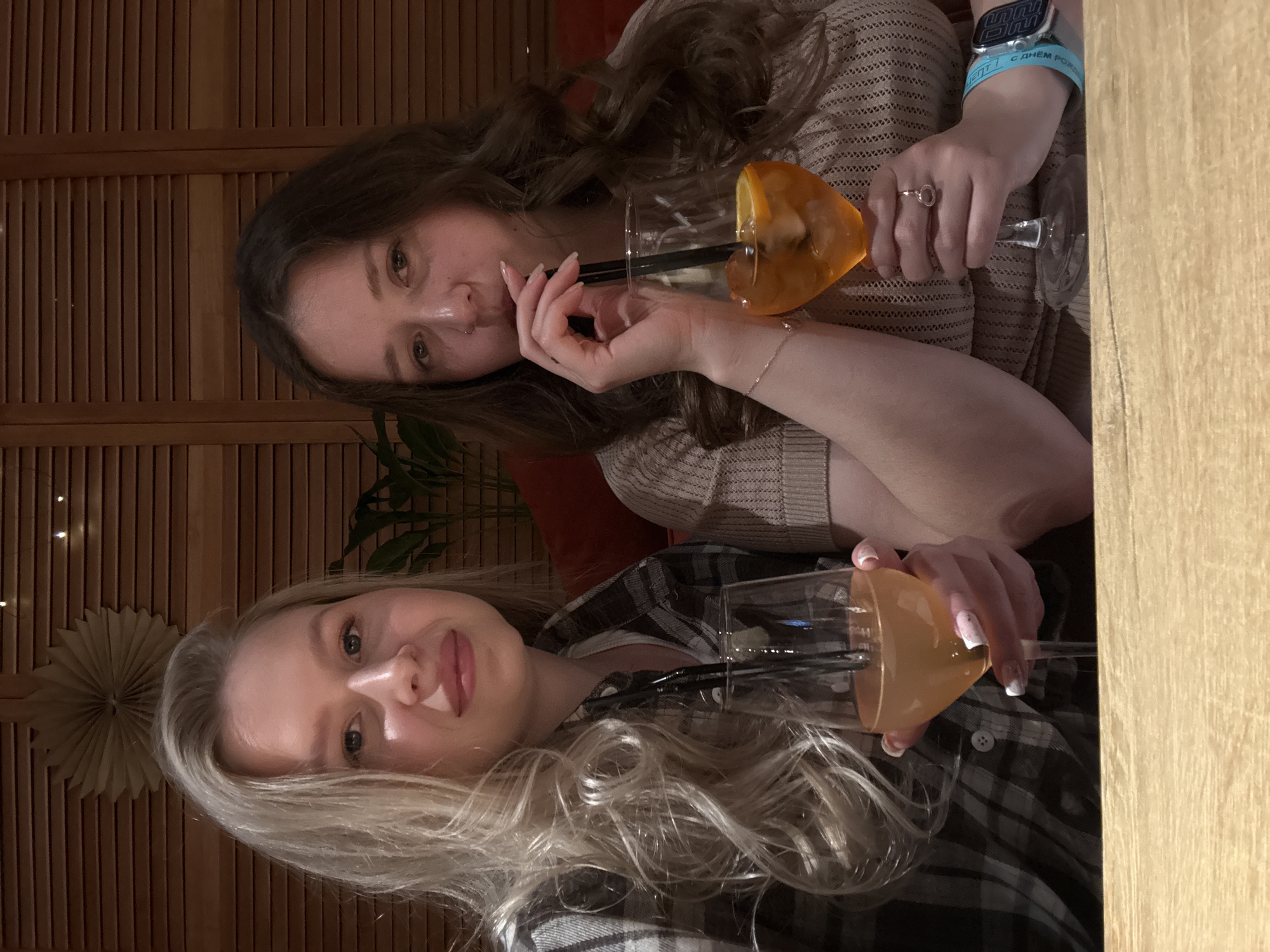 Two young women are seated indoors at a table, appearing to be socializing over drinks. The image is taken at a slight upward angle, with the subjects facing the camera.
The woman on the right has long, dark, wavy hair and is wearing a light beige or brown knitted top. She holds a stemless cocktail glass with an orange-colored drink, garnished with an orange slice, and a black straw. She is looking directly at the camera with a neutral expression, her fingers resting near her chin. She wears a ring on her right ring finger, a thin bracelet on her left wrist, and a smartwatch with a blue band. The smartwatch band has visible text that appears to be "С днём рожд..." (part of "Happy Birthday" in Russian) and "ENT".
The woman on the left has long, wavy blonde hair and is wearing a plaid shirt in shades of grey, black, and white. She also holds a stemless cocktail glass containing an orange-colored drink with a black straw. Her nails have a French tip design. She looks towards the camera with a slight smile.
The setting appears to be a cozy establishment like a bar or restaurant, characterized by dim lighting. The background features a wooden slatted wall, and a decorative paper fan or star is visible on the wall behind the blonde woman. A plant can also be seen in the background near the dark-haired woman. A wooden table surface is visible in the foreground, where both women are resting their arms. The overall mood is relaxed and social, likely taking place in the evening.