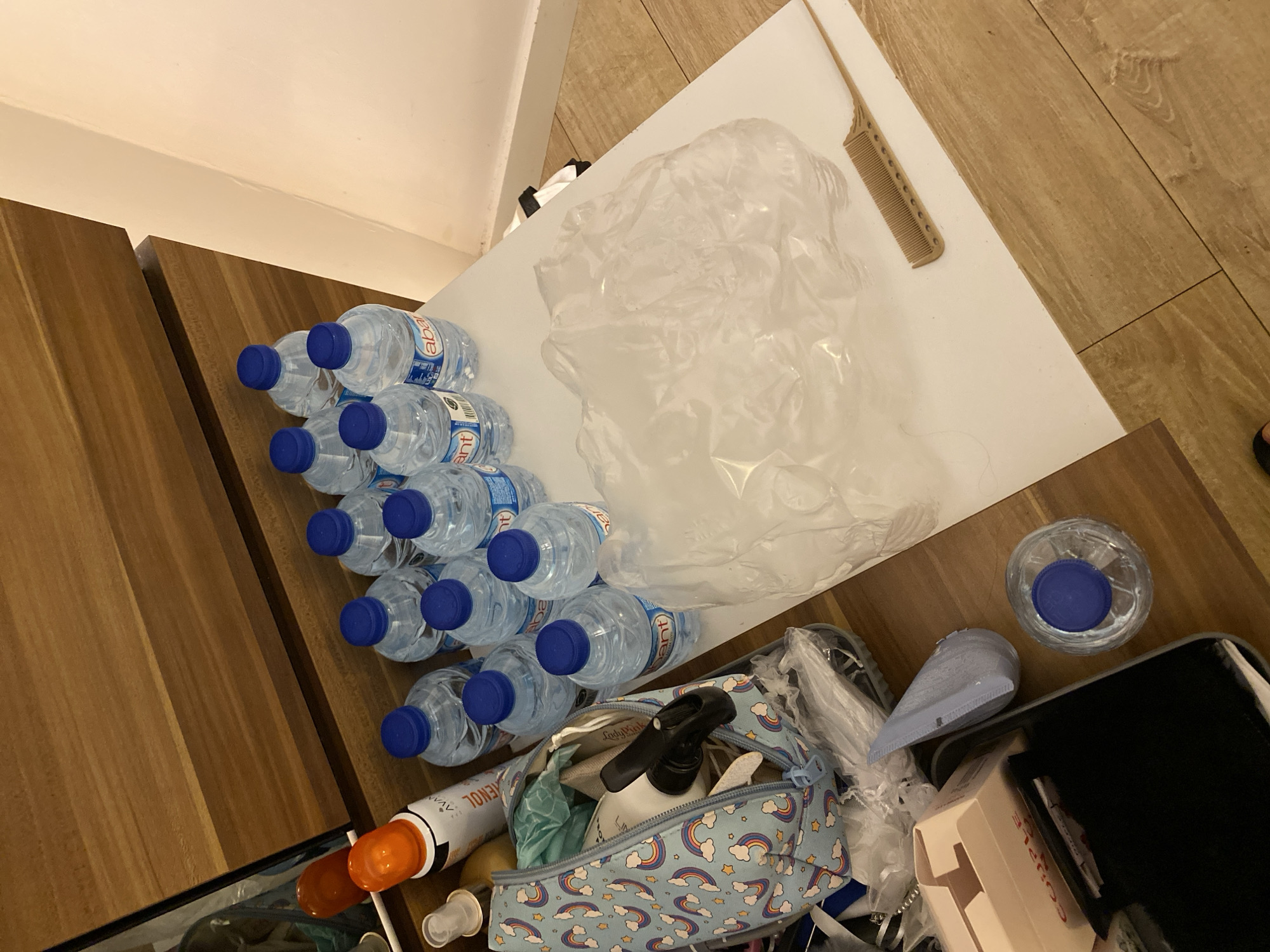 The image displays a collection of bottled water and personal items arranged on a wooden surface, likely a table or dresser. There are at least 13 plastic bottles of water, most with blue caps, organized in rows. Several other objects are present, including a cosmetic bag with a rainbow pattern, a spray can with an orange cap, a comb, and a crumpled plastic bag. A small bottle with a pump dispenser is visible inside the cosmetic bag. A single bottle of water with a blue cap is also on the wooden floor. The scene is lit by artificial light, casting shadows. The background shows a wooden cabinet and a white wall. The setting is indoors. No people are visible in the image. The presence of multiple water bottles suggests preparation for hydration or a temporary supply. The arrangement of items is haphazard, indicating a lack of specific organization. The overall scene is a still life composition of everyday objects. The image does not contain any information that could be used to identify the specific location within Aksu, Turkey.