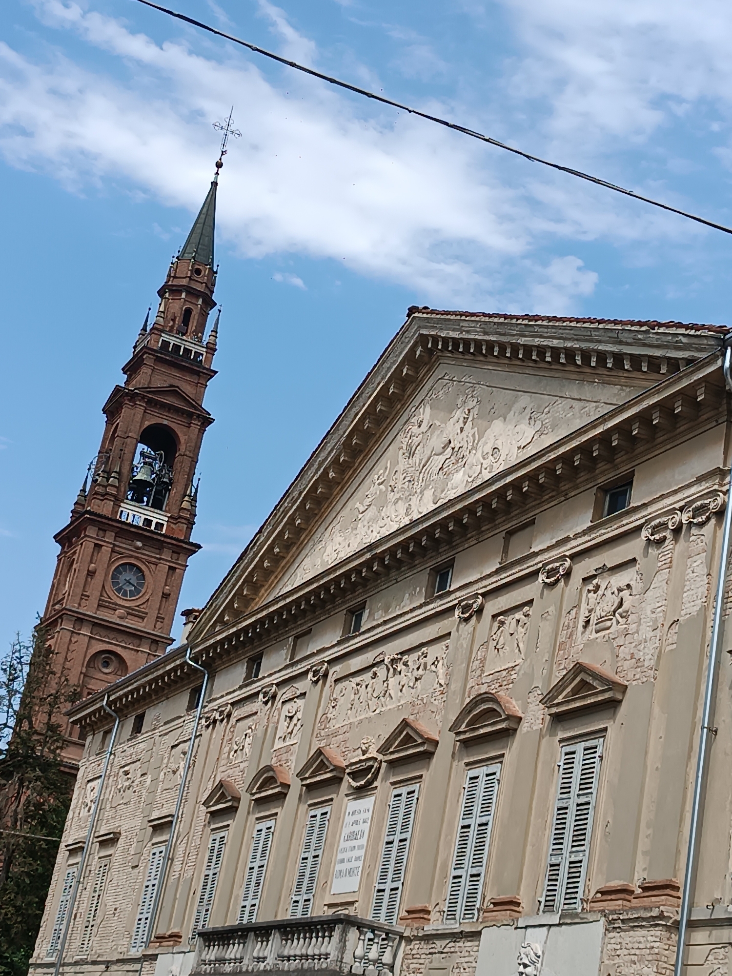 The image shows the exterior of a historic building with an ornate facade and a tall, slender bell tower. The building, located in Casalmaggiore, Italy, appears to be made of stone and brick, with visible signs of age and weathering. The facade features multiple rows of shuttered windows, decorative carvings, and a balustrade on the lower level. The bell tower, constructed from reddish-brown brick, has a clock face and a belfry containing a large bell. A spire tops the tower, adorned with a weather vane. The sky above is blue with scattered white clouds, and a power line stretches across the top of the frame. No people or animals are visible in the scene.