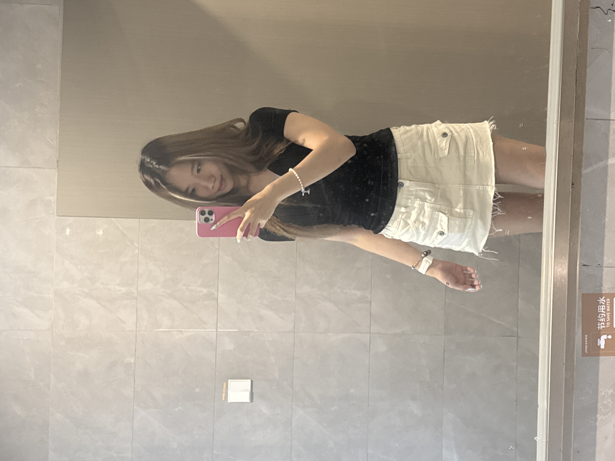 The image shows a young woman taking a selfie in a bathroom mirror. She is wearing a black top and white shorts. The bathroom has light gray tile walls. A sign in Chinese and English saying "To Save Water" is visible on the right side of the mirror. A light switch is located on the wall next to the mirror. While I can't confirm the exact location, the information suggests this could be in the Nan'an District, China.