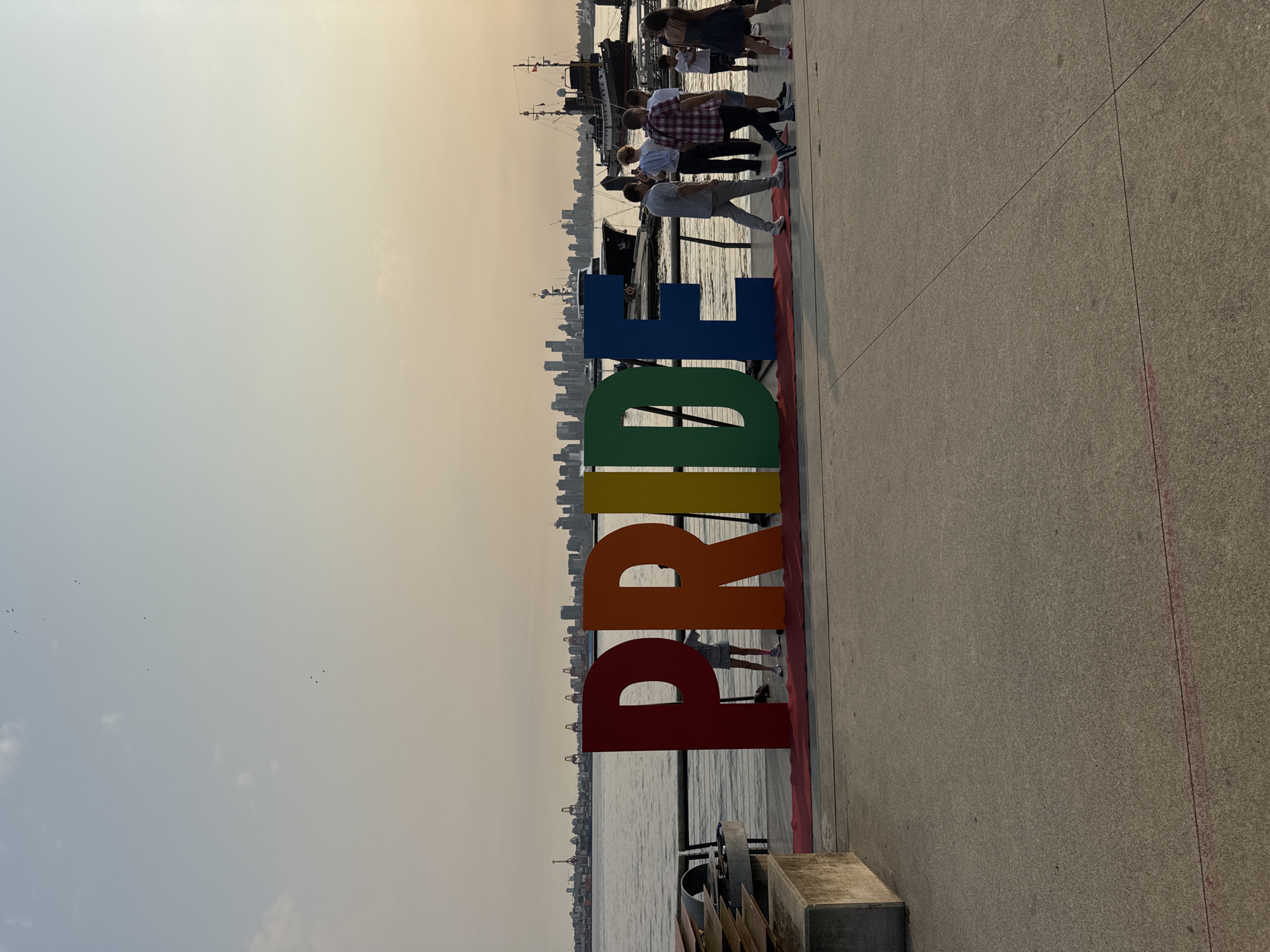 The image shows a large, colorful "PRIDE" sign on a waterfront promenade in North Vancouver, Canada. The letters are red, orange, yellow, green, and blue. Several people are walking around the sign. In the background, a city skyline with many buildings and cranes is visible across the water. Two boats are docked in the water near the promenade. The sky is overcast and the sun is setting, casting a warm glow over the scene.
