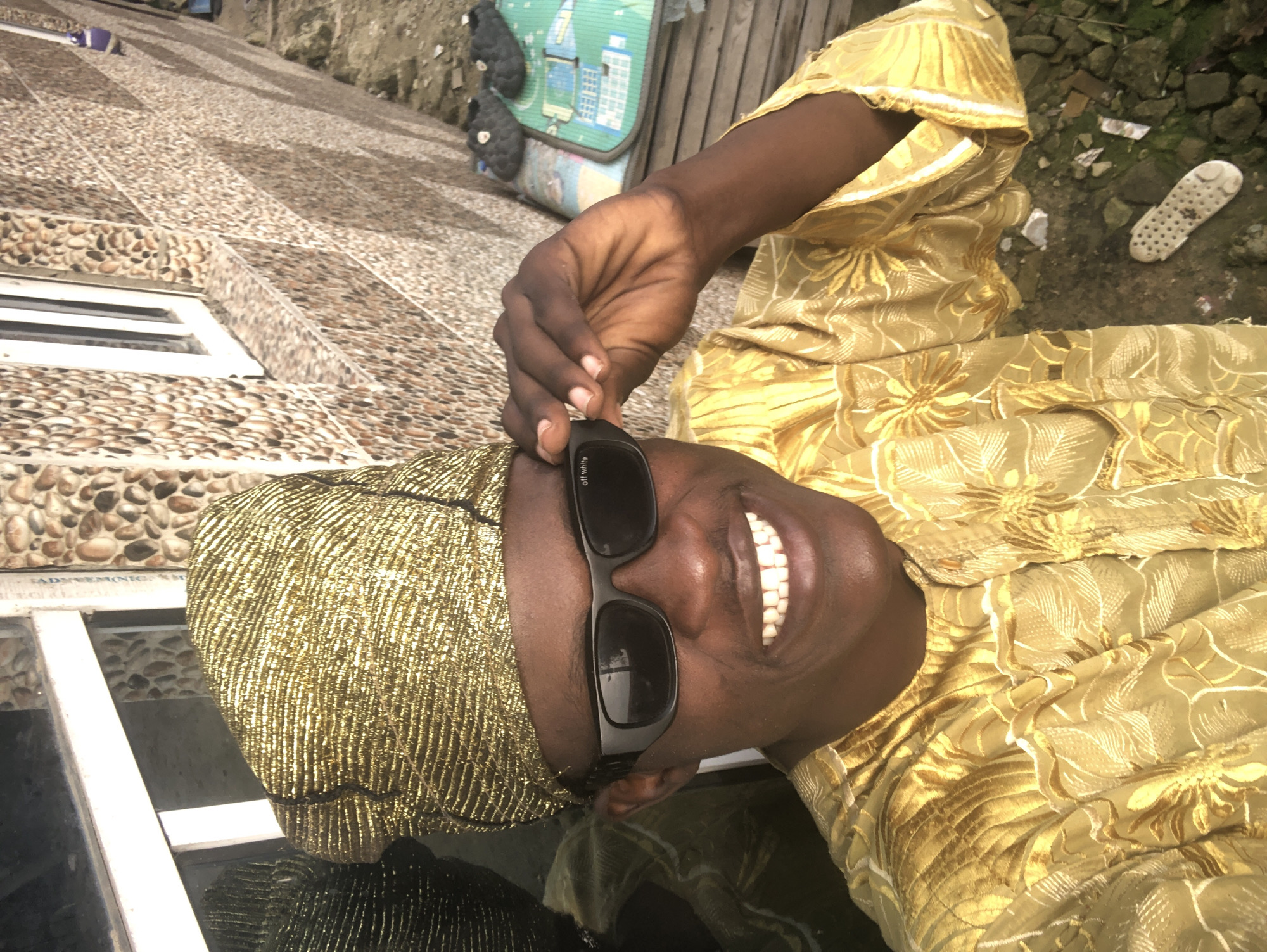 In Ado, Nigeria, a man in a gold brocade outfit and matching headwrap smiles broadly for a selfie. He wears black sunglasses, and his left hand adjusts them. The man's attire features intricate floral embroidery in gold thread. Behind him, a wall is decorated with a mosaic of rounded stones. A white-framed window with a partial reflection of the man is visible. The ground is covered with rocks and debris, and a green mat with a blue border lies on the ground, topped with a pair of dark slippers. A single white sandal with a perforated sole is also visible on the ground.