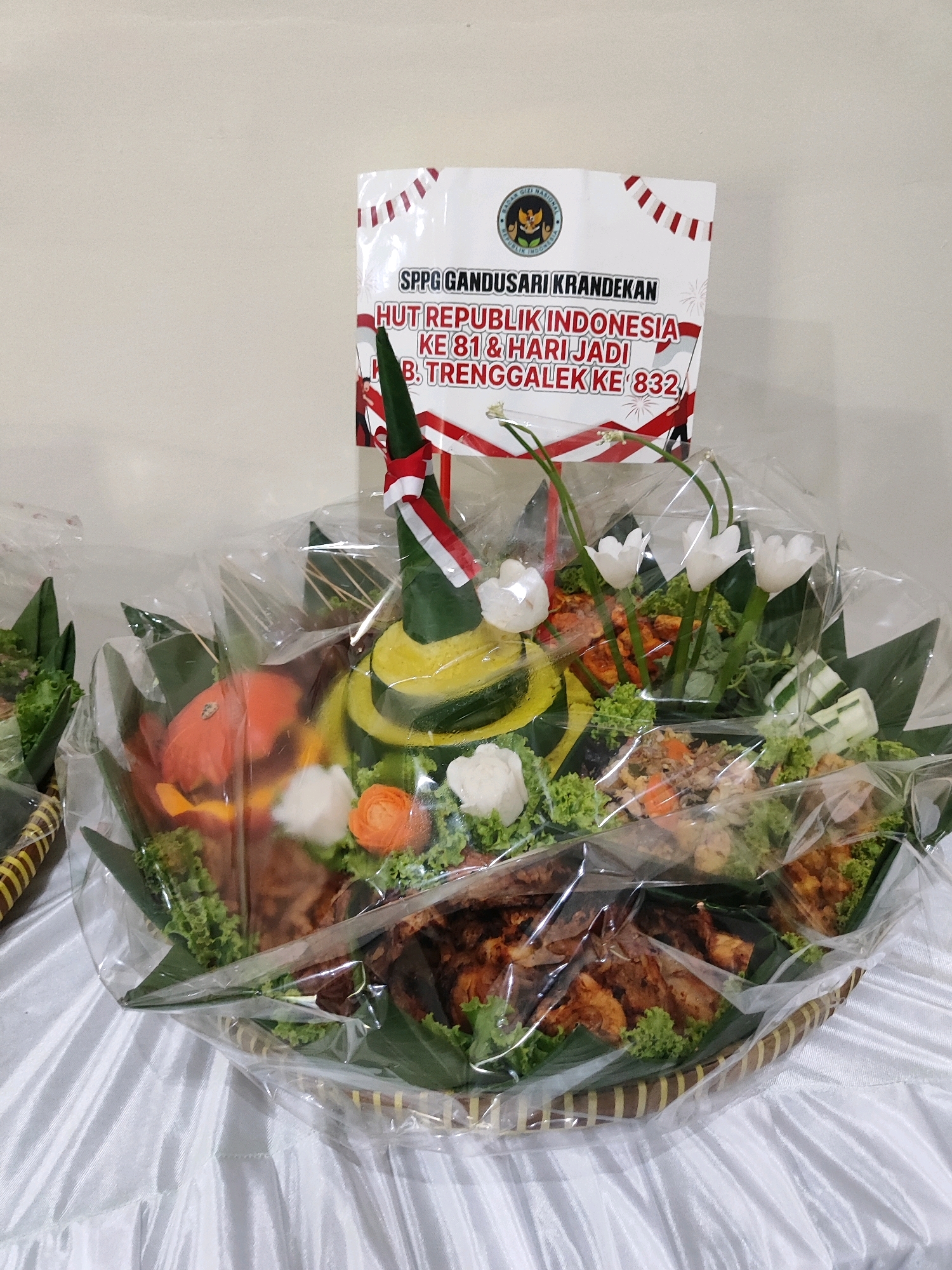 The image shows a large, elaborately decorated food platter, likely a traditional Indonesian tumpeng, presented on a white tablecloth. The tumpeng is conical in shape and covered in green and yellow layers, adorned with carved vegetables and white flowers. The platter itself is made of woven material and is covered in clear plastic wrap.
A white banner is placed behind the tumpeng, featuring Indonesian text and a national emblem. The text on the banner reads "SPPG GANDUSARI KRANDEKAN HUT REPUBLIK INDONESIA KE 81 & HARI JADI B. TRENGGALEK KE 832". This indicates that the tumpeng is a celebratory offering for the 81st anniversary of the Republic of Indonesia and the 832nd anniversary of the Regency of Trenggalek. The city of Trenggalek is located in East Java, Indonesia.
The overall scene suggests a formal occasion or celebration, possibly a cultural event or a departmental gathering within the Gandusari Krandekan area. The lighting appears to be indoor, with a soft, diffused light source. There are no people visible in the image.