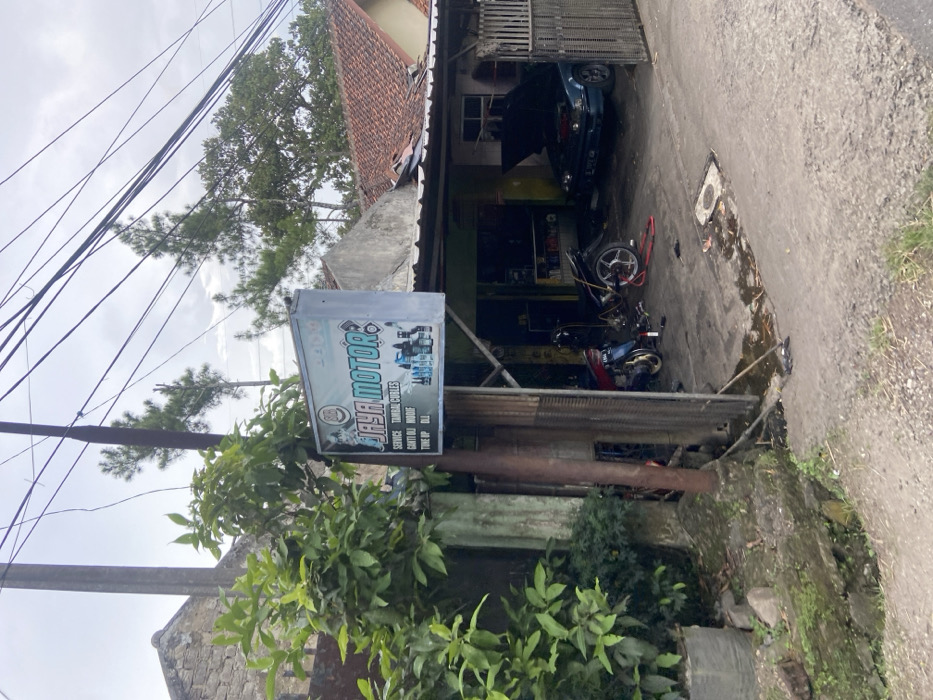 The image shows a small auto repair shop in Cipatat, Indonesia, located at latitude -6.787 and longitude 107.434. The shop has a sign that reads "Jaya Motor" in large blue lettering with a black outline. The sign lists several services, including "Service," "Tambal Cules," "Ganti Oli," "Modif," and "Tune Up Oli." The shop is located on a narrow street with a small concrete area in front of the shop where vehicles are parked. The shop has a partially open garage door, and there are several vehicles inside, including a black motorcycle with a silver exhaust pipe, a blue car with the hood open and some tools lying nearby, and a blue car with a yellow license plate. The shop is located on a side street with a lot of vegetation growing nearby. There are several power lines running overhead. The sky is overcast and gray. The ground is covered with dirt, rocks, and vegetation. The image was likely taken from the street.