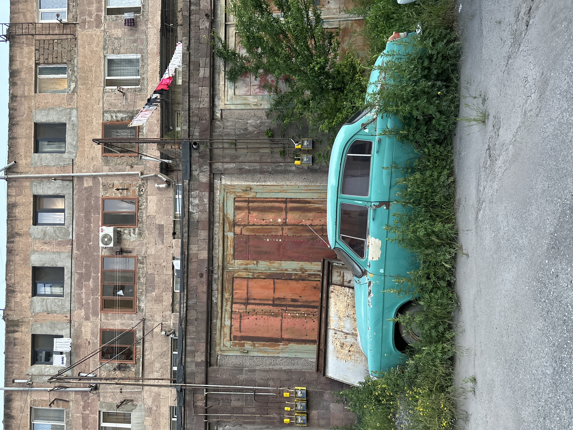 The image shows an abandoned blue car overgrown with vegetation, parked next to a weathered building in Yerevan, Armenia. The building has multiple windows, some with air conditioning units, and clothes hanging to dry on a line extending from one of the upper floors. A large, rusted metal door is visible at street level of the building. Utility meters are mounted on the wall near the base of the building. The scene is set on a cracked asphalt street.