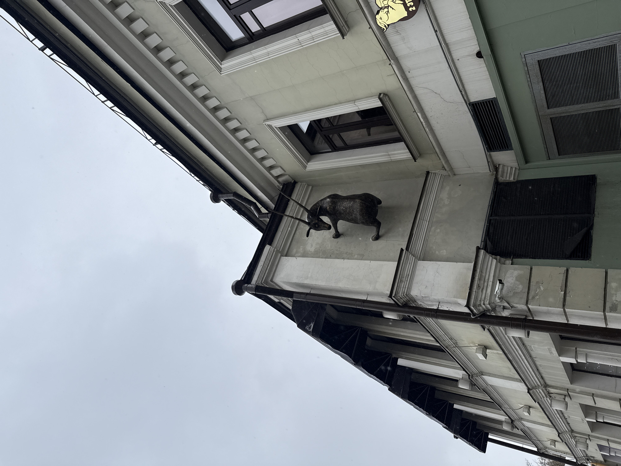 A low-angle shot shows the facade of a building. The building is light-colored and has decorative architectural details, including cornices and window frames. There's a sculpture of an animal, possibly a deer or antelope, mounted on the exterior wall. The sky is overcast and white. From the image, it can be deduced the building is in Moscow, Russia, due to the presence of a sign in Russian with the name "2 этаж".