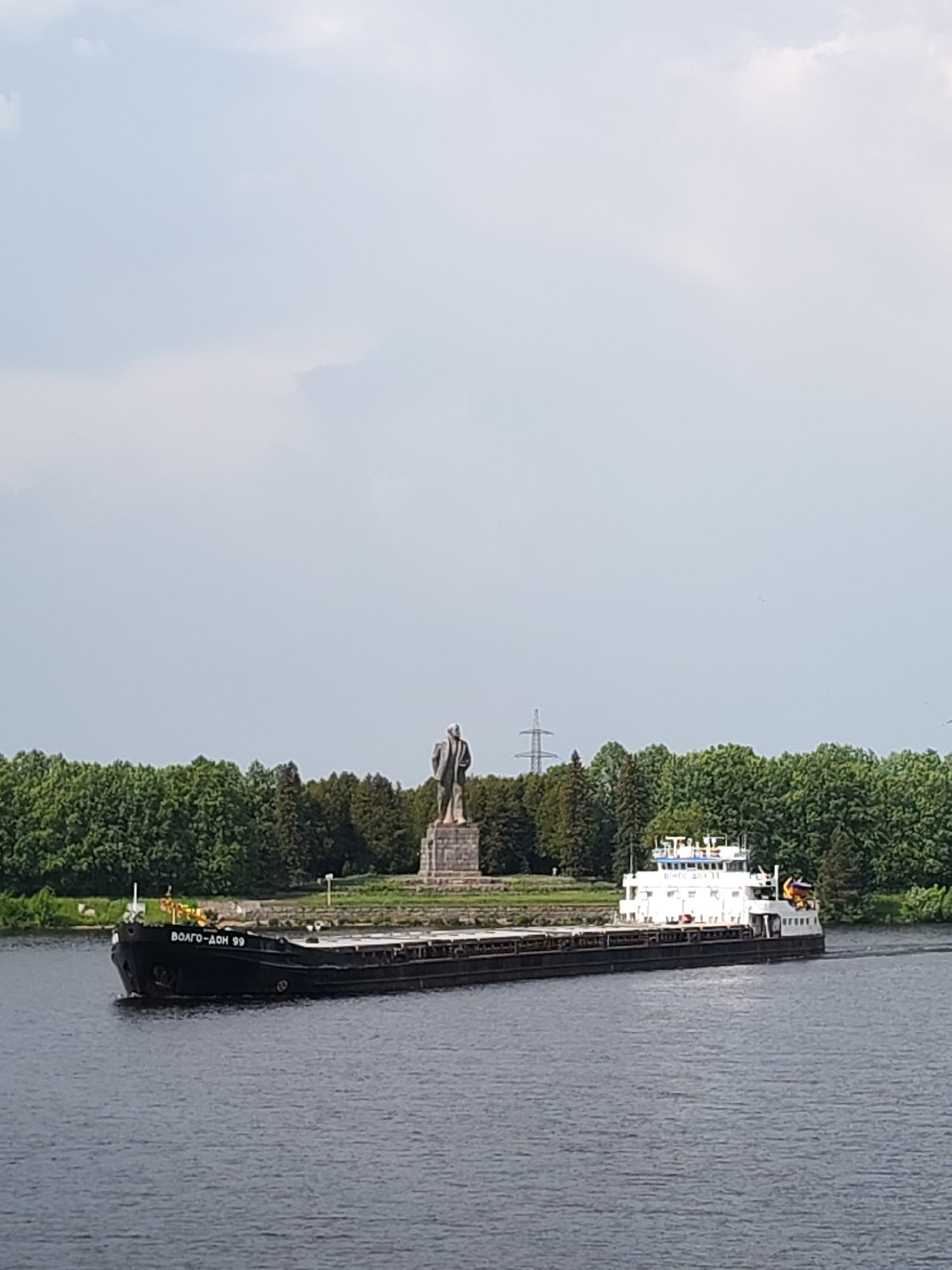A barge is sailing on a river in front of a statue. The barge is black and white and has the name "VOLGO-DON 99" painted on the front. The statue is of a man in a suit and tie, and it is standing on a pedestal in a park. The park is surrounded by trees. The sky is overcast. The location appears to be near the Volga–Don Canal.