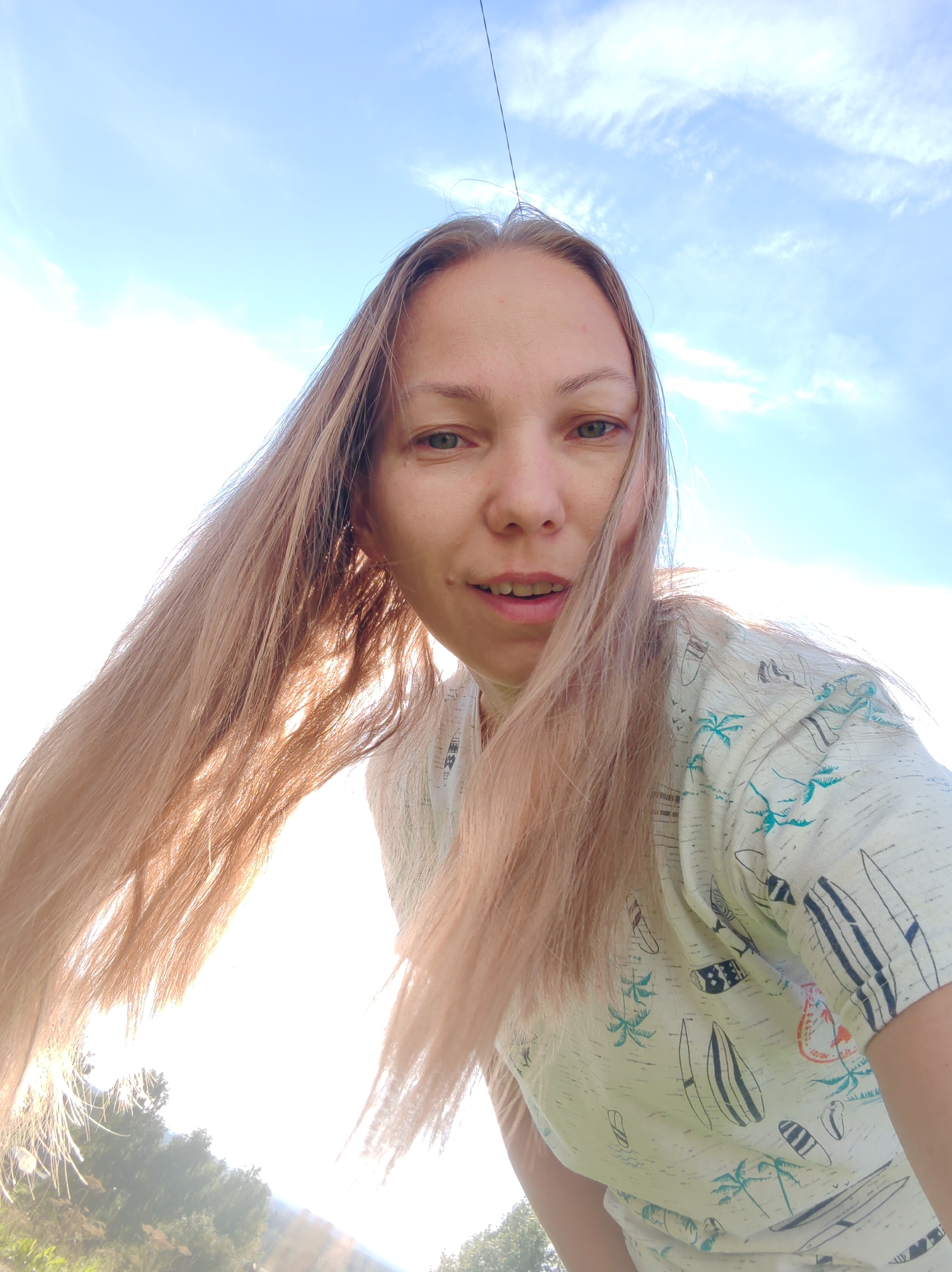A young woman with long, blonde hair is taking a selfie outdoors. She is wearing a light-colored t-shirt with a tropical print featuring palm trees and surfboards. Her hair is blowing to the side, and the sky is visible behind her, with scattered clouds. The lighting suggests it is a sunny day. The background shows hints of greenery and trees. The image is taken from a low angle, looking up at the woman. The location is not discernible from the image.