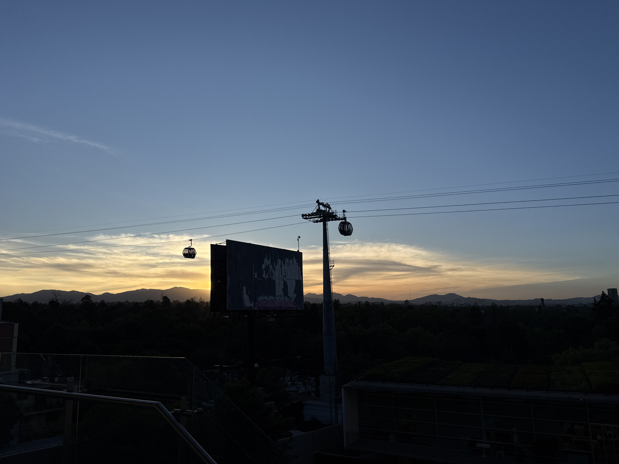 The image captures a view in Mexico City, Mexico at dusk. The sky is a gradient of blue and orange, indicating either sunrise or sunset. Silhouetted against the sky are mountains and buildings in the distance. In the foreground, there's a cable car system with two gondolas and a support tower, all in silhouette. Behind one of the gondolas, a large billboard blocks the sun, also silhouetted. Below, there is a dark expanse of trees with some buildings visible near the bottom edge of the frame. A railing and some elements of the building where the photo was taken are visible in the bottom portion of the image.