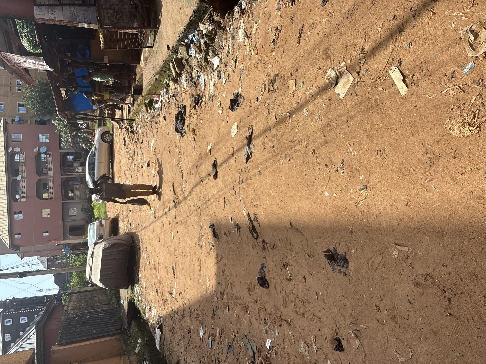 In Nkpor, Nigeria, a dirt road is littered with trash, including plastic bags, wrappers, and other debris. A man walks down the road carrying a bag over his shoulder. In the background, a car is covered with a tarp, and several buildings are visible. A small market stall is set up on the side of the road, with a few people standing around it. The overall scene depicts a busy, urban environment with visible signs of litter and disorganization.