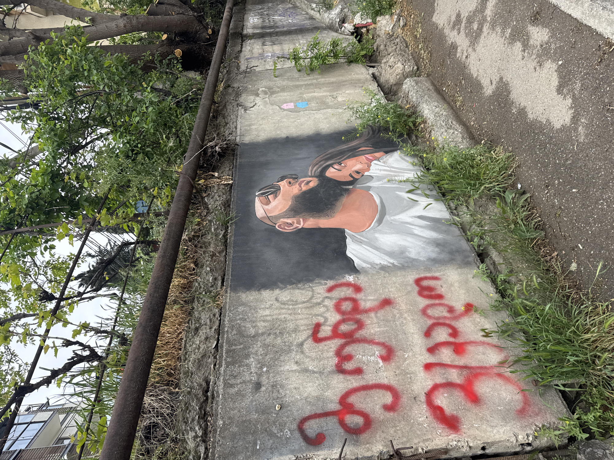 The image shows a concrete retaining wall in Tbilisi, Georgia, with a mural of a man and a woman painted on it. The man has a beard and is wearing sunglasses. The woman has long hair and is looking up. Underneath the mural, there is red graffiti. There are trees and plants growing near the wall, and a sidewalk next to it. A rusty metal pipe runs along the top of the wall. The overall impression is one of urban decay and street art.