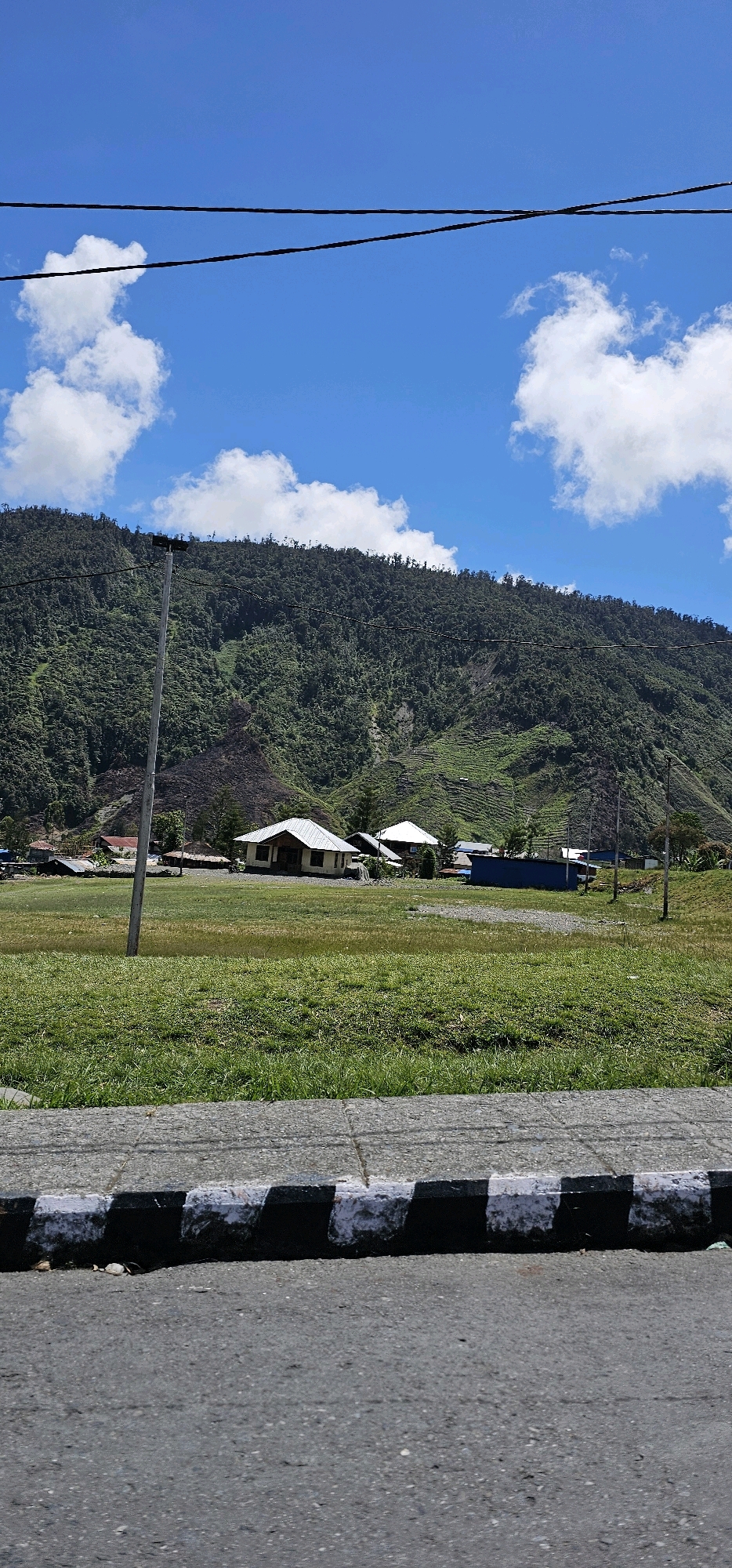 The image depicts a rural mountainous landscape under a bright blue sky with scattered white clouds. In the foreground, a paved road is visible with a black and white striped curb. Beyond the curb, a grassy field stretches towards a cluster of buildings, including houses with light-colored roofs and a blue structure. A utility pole stands in the middle of the field. The background is dominated by a lush green, densely forested mountain slope, with some areas showing signs of erosion or agricultural terracing. The overall scene suggests a remote village or settlement nestled at the foot of a mountain in a tropical or subtropical region.