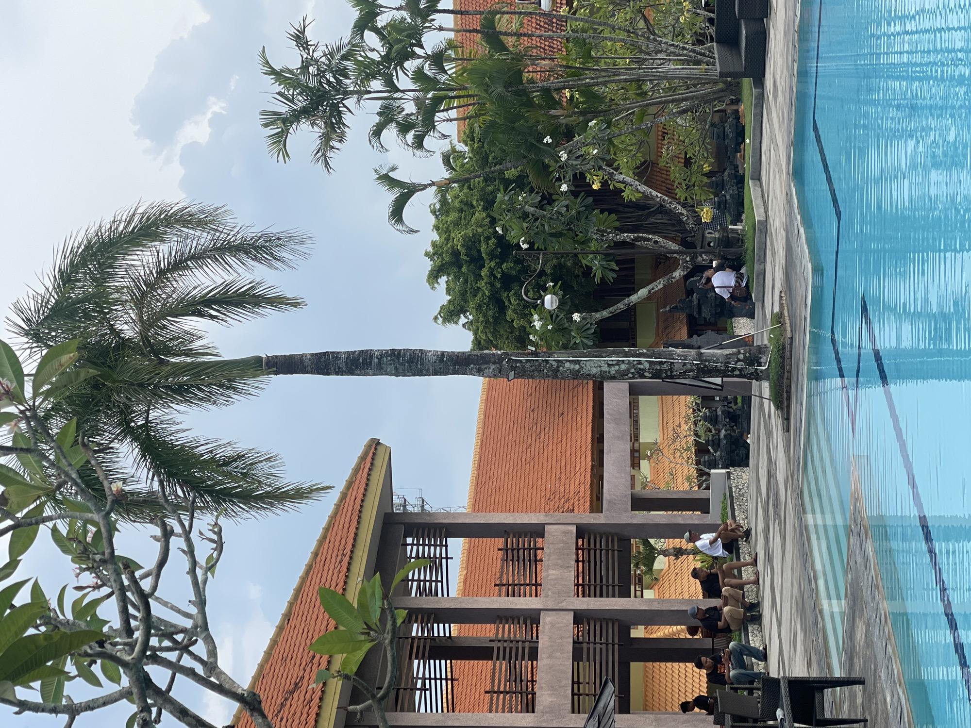 In Kuta Selatan, Indonesia, an outdoor scene features a swimming pool with bright blue water in the foreground. Several individuals are seated on a stone bench to the left of the pool, facing the water. Behind them, a multi-story building with red tile roofs and brown columns is visible. A tall palm tree stands prominently in the center of the image, with other tropical foliage and smaller palm trees to the right. The sky is overcast with clouds. The overall environment appears to be a resort or hotel complex.