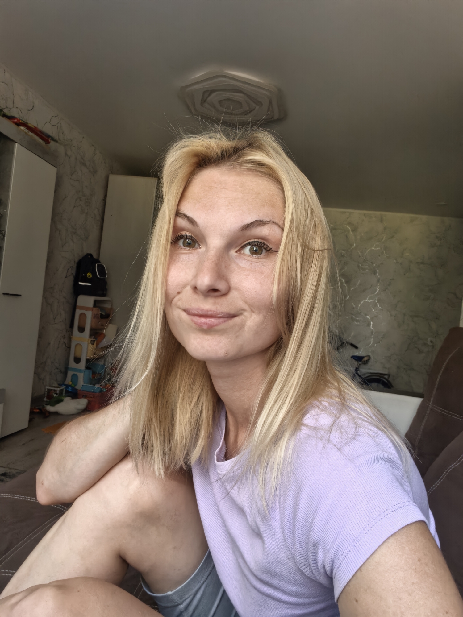 A blonde woman with light skin and freckles is sitting on a brown couch, looking at the camera. She is wearing a light purple t-shirt and gray shorts. Her right arm is bent at the elbow and resting on her lap. Her left arm is bent and resting on her leg. In the background, there is a white wardrobe, a backpack on a shelf, and various children's toys scattered on the floor. The walls have a marble-like pattern. The room appears to be a living room in an apartment. The lighting suggests it is daytime. The image was taken indoors.