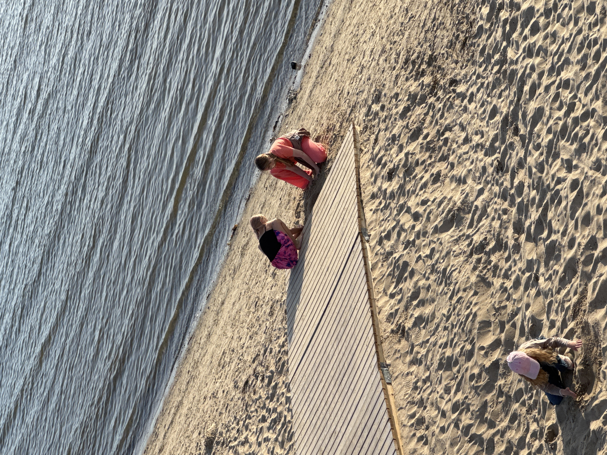 Three young people are on a sandy beach next to a body of water, likely the Baltic Sea, in Tallinn, Estonia. The two individuals closest to the water are crouching near a wooden boardwalk that is partially submerged in the sand. The person on the left wears a black top and a pink and purple patterned skirt. The person on the right wears an orange top and pants, with a brown bag slung over their shoulder. Further up the beach, a third individual wearing a pink cap, a beige top, and dark pants is also crouching and interacting with the sand. The water is calm with small ripples, and the beach sand shows indentations from footprints and wind patterns. The lighting suggests it is late afternoon or early evening.