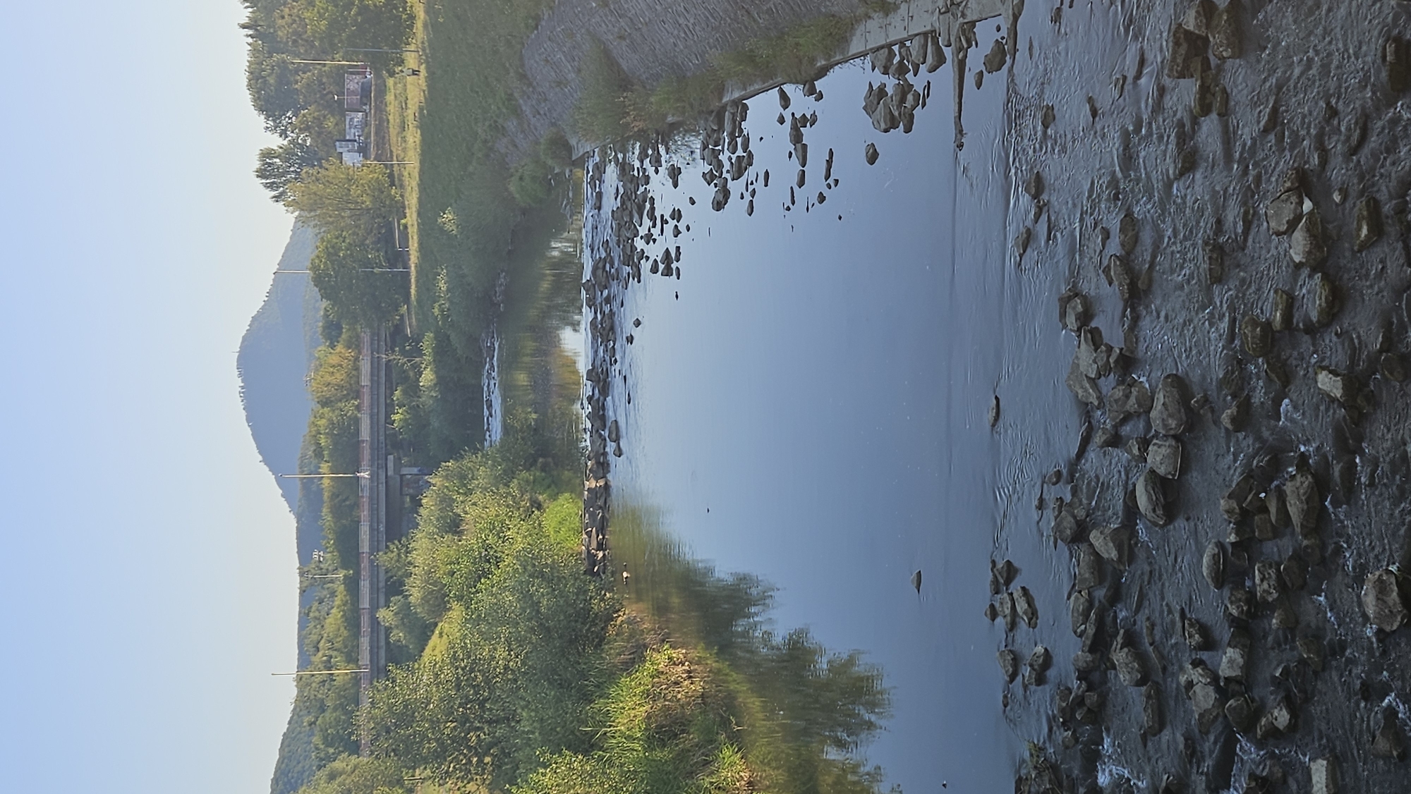 The image shows a view of the Torysa River flowing through the city of Prešov, Slovakia. The river is shallow with visible rocks and rapids in the foreground. The water is calm and reflects the blue sky and surrounding greenery.
On the right bank, a concrete embankment lined with stones is visible. On the left bank, lush green vegetation and trees line the river. In the background, a bridge spans the river, with trees and hills beyond. A prominent, rounded hill with a structure on its summit is visible in the distance, dominating the skyline. The overall scene suggests a peaceful, natural environment within an urban setting, likely during daylight hours with clear weather. There are no people or animals clearly visible in the image.