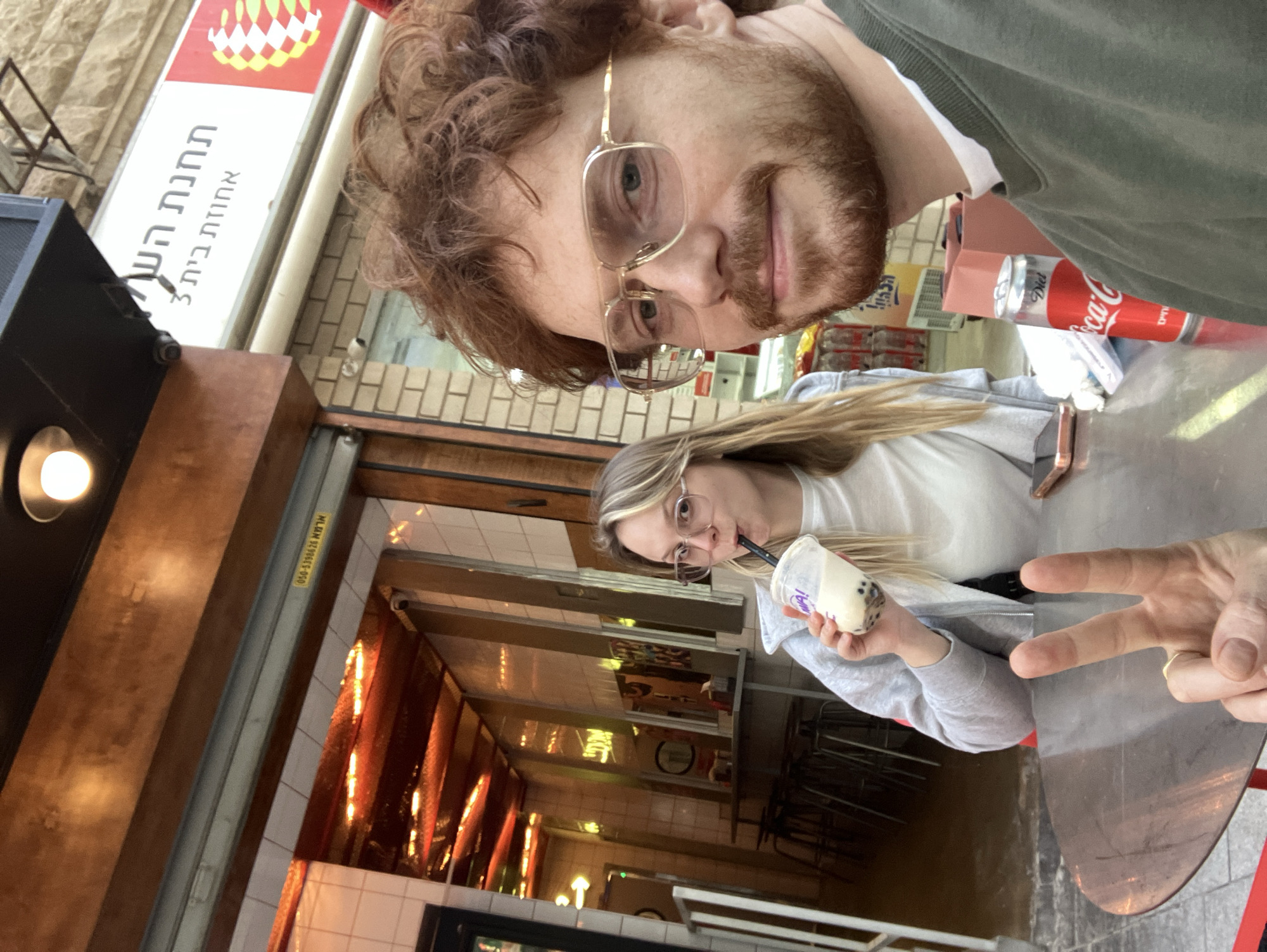 A medium, slightly angled selfie depicts a man with reddish-brown hair and beard, wearing glasses and an olive green jacket, looking directly at the camera. Behind him, slightly out of focus, a woman with blonde hair, also wearing glasses, is drinking from a cup with a straw. A hand in the foreground makes a peace sign. They are sitting at an outdoor table.
Behind them is the storefront of a restaurant. A sign in Hebrew is visible, but not legible. The restaurant interior features a long, narrow space with warm lighting and tile walls. Canned drinks are visible behind the table. A sticker on the door indicates the location as Tel Aviv, Israel.