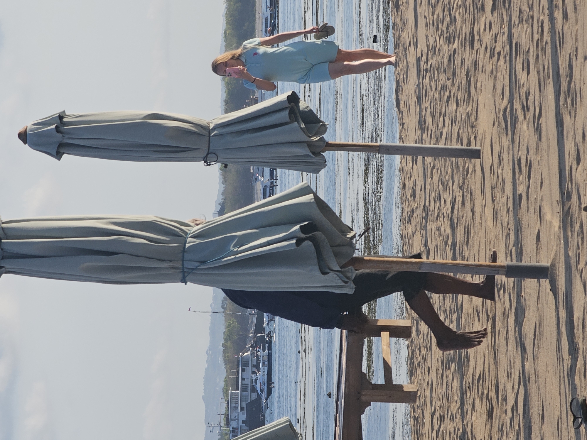 On a sandy beach in Denpasar, Indonesia, two large, closed beach umbrellas stand in the foreground. A person in a dark blue top and dark shorts walks behind one of the umbrellas, with their body partially obscured by the fabric. Their left leg is extended forward in a walking motion, and their bare foot is visible. In the background, a woman in a light blue romper is walking to the right, holding a pink smartphone in her right hand and a pair of flip-flops in her left. She is looking at her phone. The sea is visible behind the beach, with several boats and a distant, hilly coastline.