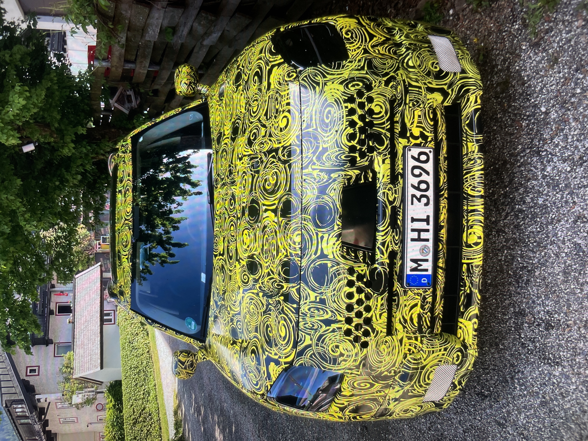 The image shows a car with a black and yellow swirly camouflage pattern. The car is parked on a paved road next to a hedge. The license plate reads "M HI 3696" and has a German flag on it. In the background, there is a house and some trees. The city is unknown.