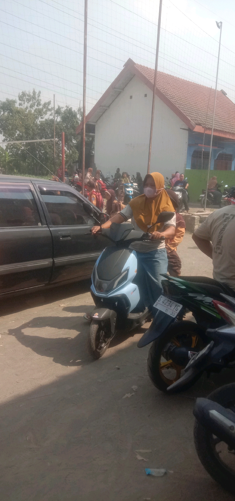 In Cangkringrandu, Indonesia, a woman wearing a mustard-colored hijab and a pink face mask rides a light blue scooter. She is wearing blue jeans and her right hand is on the scooter's handlebar. Behind her, a person wearing a brown and orange shirt is seated on the back of the scooter. To her left, a dark-colored car is parked. In the background, other people are visible, many of whom appear to be on motorcycles, and a white building with a red tiled roof stands under a bright, overcast sky. The scene appears to be an outdoor gathering or waiting area, possibly near a school or community center, given the presence of multiple motorcycles and people. The ground is a paved surface with some scattered debris.
