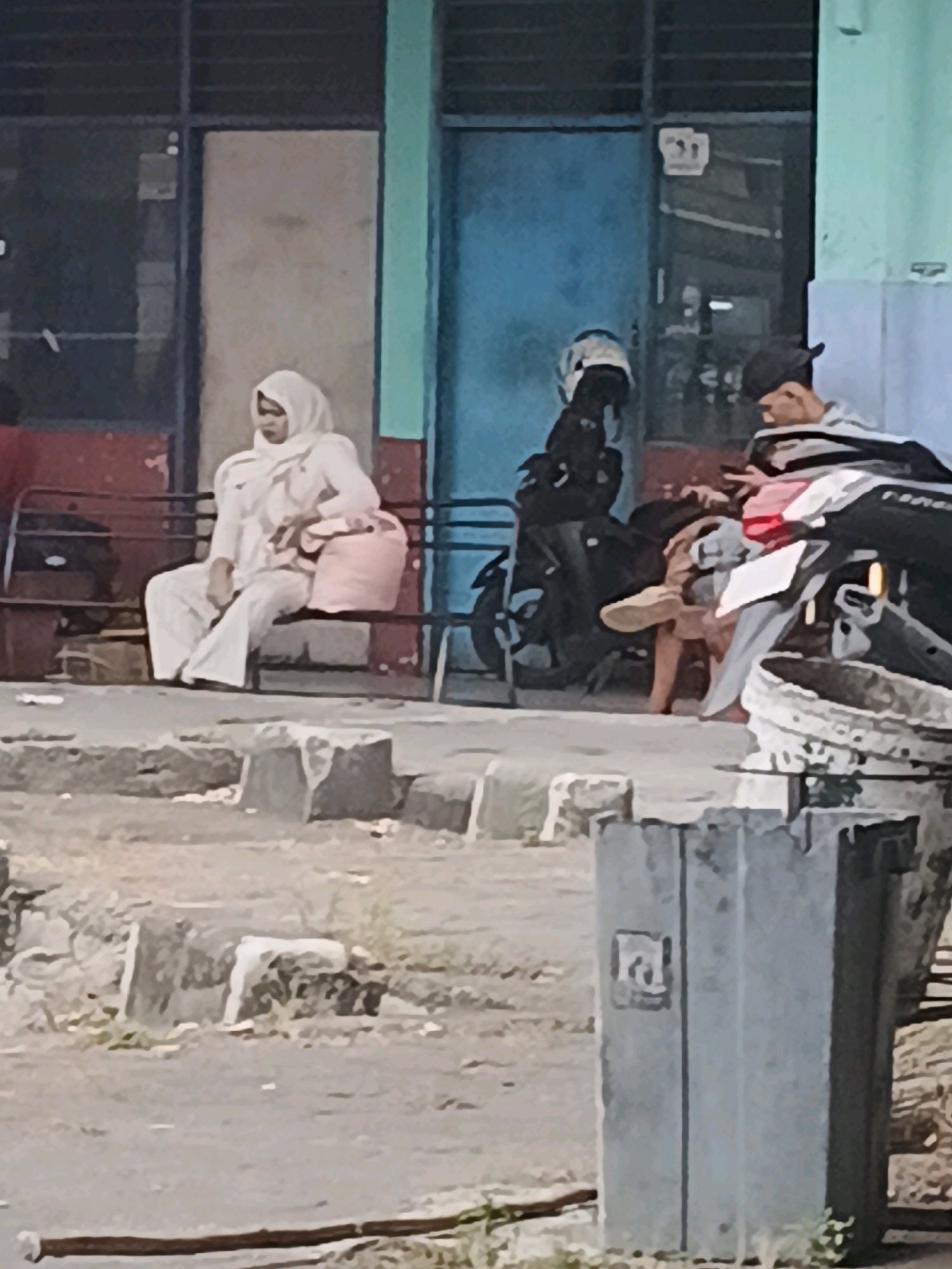 The image depicts a street scene in the city of Kuripan, Indonesia. In the foreground, a person wearing a white hijab and light-colored clothing sits on a dark metal bench, holding a pink bag. To their right, another individual wearing a dark baseball cap and a striped shirt is partially visible, appearing to interact with a smartphone. Behind them, a black motorcycle with a white and blue helmet is parked. The background features a building with blue and teal sections, and some windows. The ground appears to be paved with rough surfaces and scattered debris. A concrete trash receptacle stands in the lower right corner of the frame.