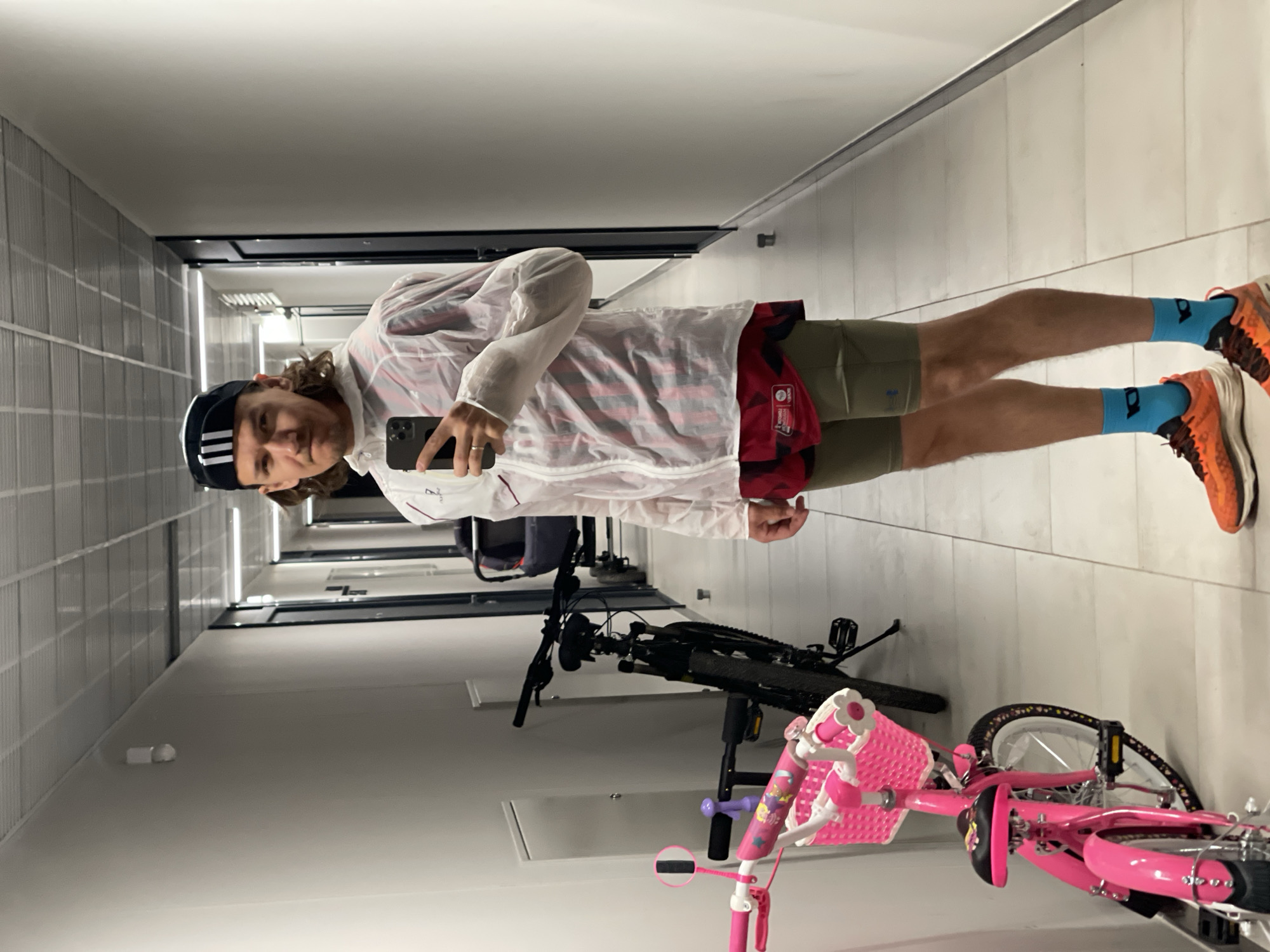 A person stands in a narrow, brightly lit corridor, taking a selfie with a smartphone. The corridor has a row of doors on the left and a tiled wall on the right. The person is wearing a dark cap with white stripes, a semi-transparent white jacket over a red and black plaid shirt, olive shorts, blue socks with black designs, and orange running shoes. A black mountain bike and a pink child's bicycle are partially visible in the lower portion of the image. The location is not identifiable as Izhevsk, Russia.