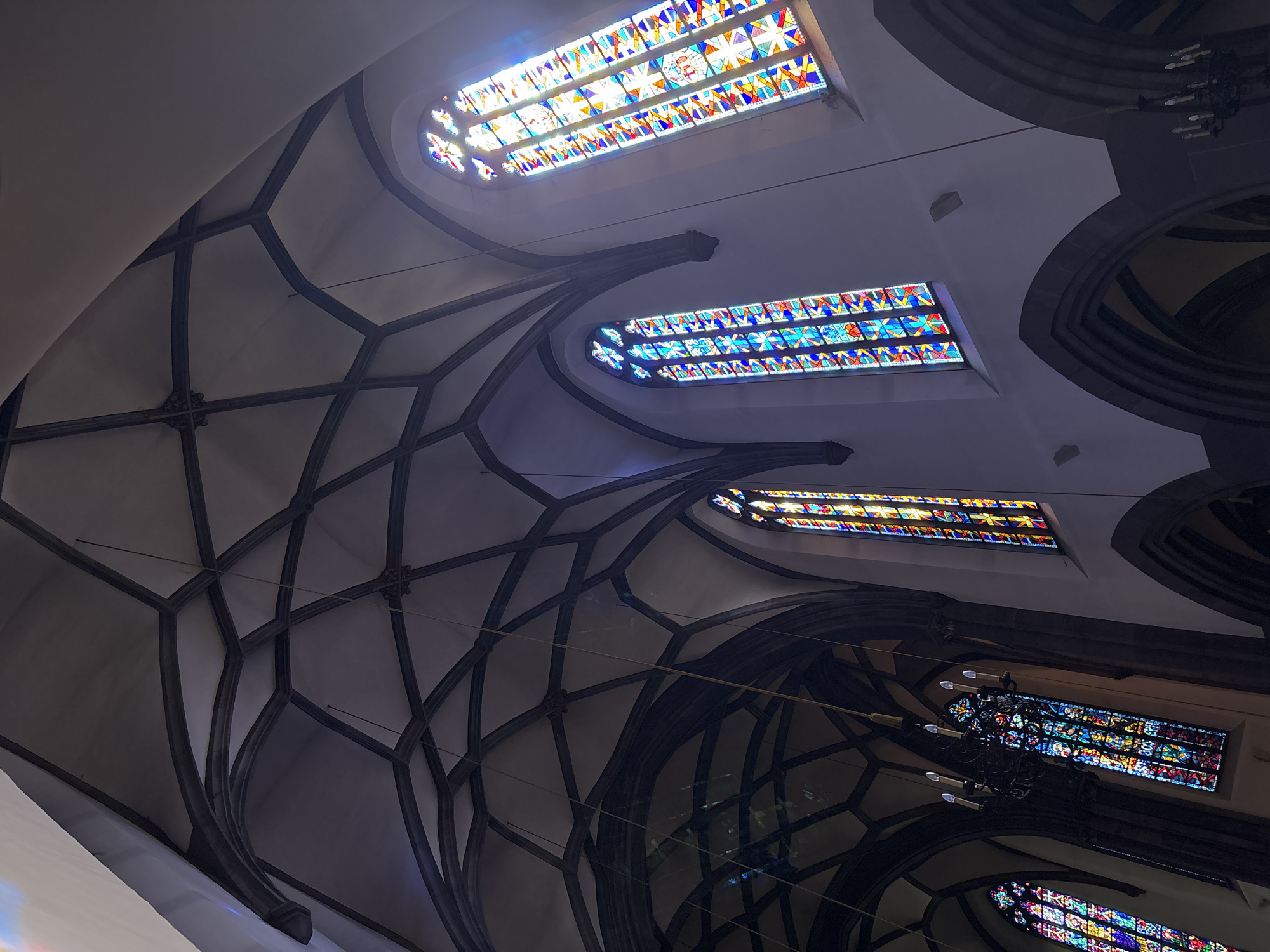 The image shows the interior of a cathedral in Strasbourg, France, specifically the Strasbourg Cathedral. The scene is dominated by a high, vaulted ceiling with Gothic architecture, characterized by dark, ribbed vaults. Sunlight streams through large, ornate stained-glass windows, casting colorful patterns of light onto the white walls and the ceiling. Three prominent stained-glass windows are visible, each featuring intricate geometric designs and vibrant colors like blue, red, yellow, and white. One window is centrally located on the right wall, a second is on the left wall further in the background, and a third is visible at the far left edge of the frame, partially obscured by the architecture. Hanging from the ceiling are at least two chandeliers, one dark and metallic with multiple candle-like lights, and another less distinct one further back. The arches of the cathedral's structure are also visible, with dark stone contrasting with the lighter walls. The overall impression is one of grandeur, history, and spiritual illumination.