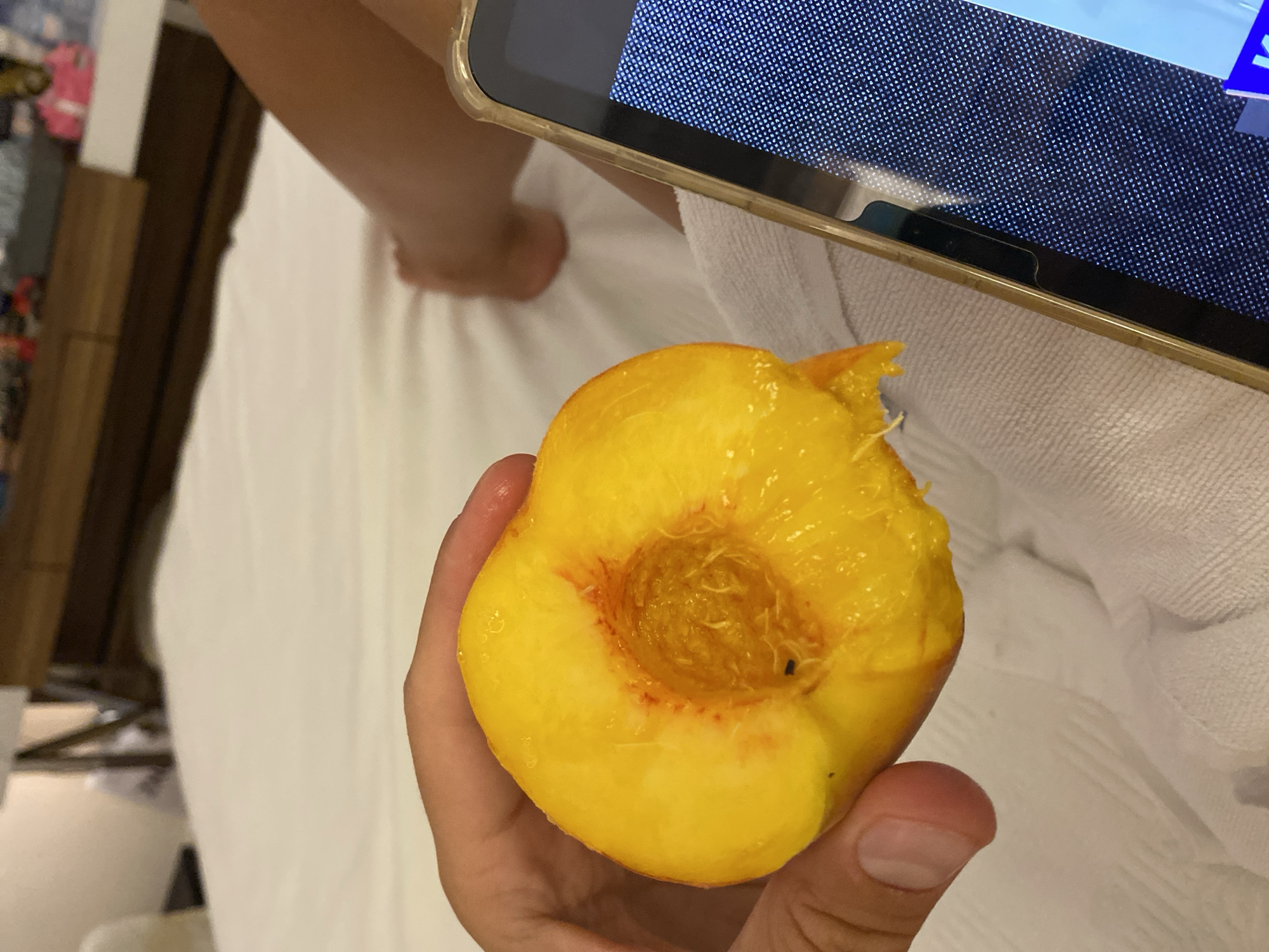 An extreme close-up shot shows a hand holding a halved peach over a white towel on a bed. The peach is bright yellow with red blushing near the pit, and it appears juicy. To the right of the peach, a portion of a smartphone is visible, displaying a textured blue screen. In the background, out of focus, are a person's legs and feet, and a dresser with items on top. The scene suggests a relaxed, indoor setting. There is no information that indicates the location is Aksu, Turkey.