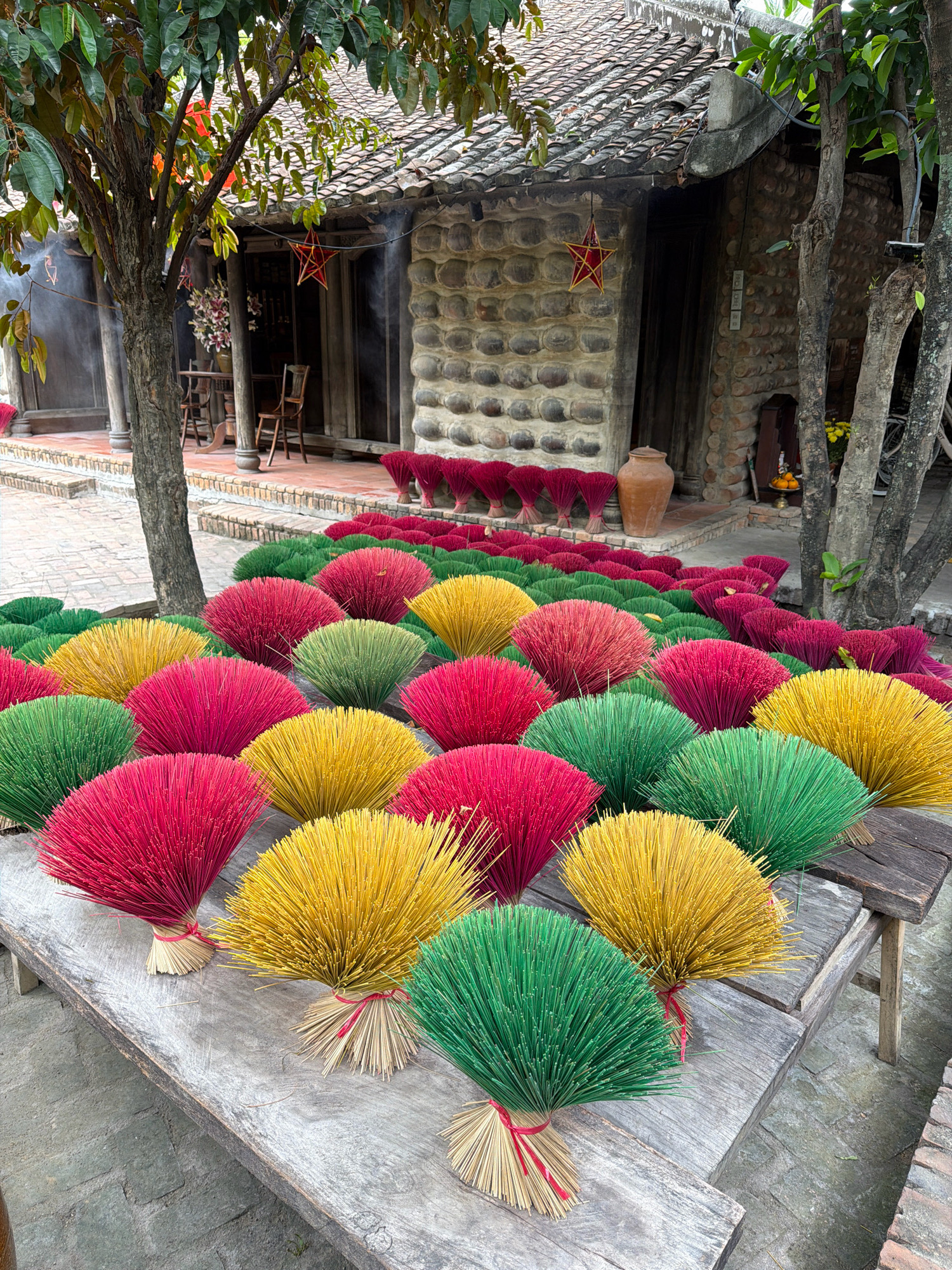 In Nha Trang, Vietnam, a vibrant display of incense sticks is arranged in colorful bunches on a weathered wooden table. The sticks are predominantly red, yellow, and green, fanned out in semicircles. Behind the table, a traditional Vietnamese house with a tiled roof and stone walls stands as the backdrop. The house has an open entrance, revealing a glimpse of furniture and decorations inside, including a red star ornament hanging from the eaves. Trees with lush green leaves frame the scene, and smoke is visible, possibly from incense being burned. The ground is paved with bricks.